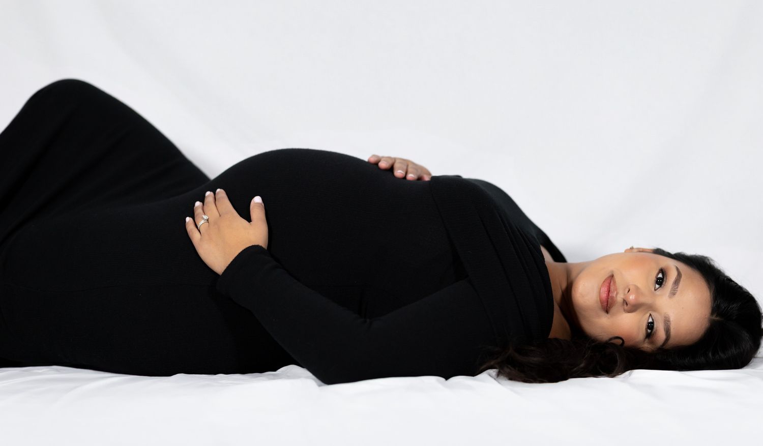 A pregnant woman in a black outfit lies on her back on a white background creating an artistic fashion pose.
