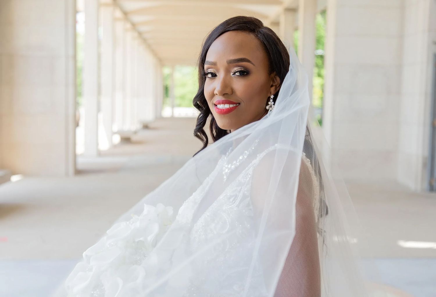 Radiant bride in white veil poses in sunlit colonnade.