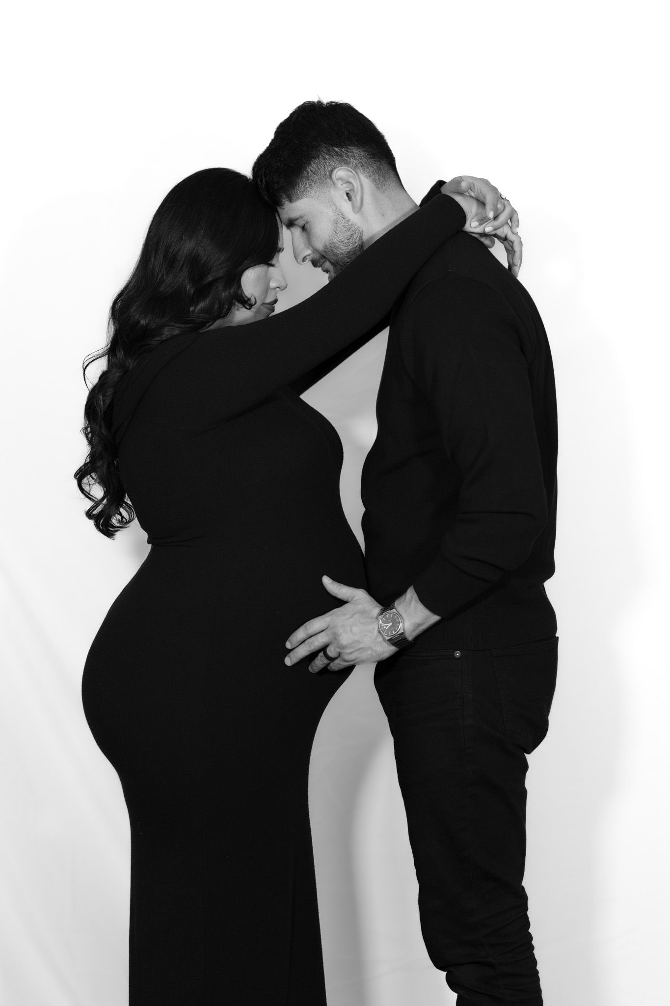 Black and white maternity photo of couple embracing in intimate pose against white background.