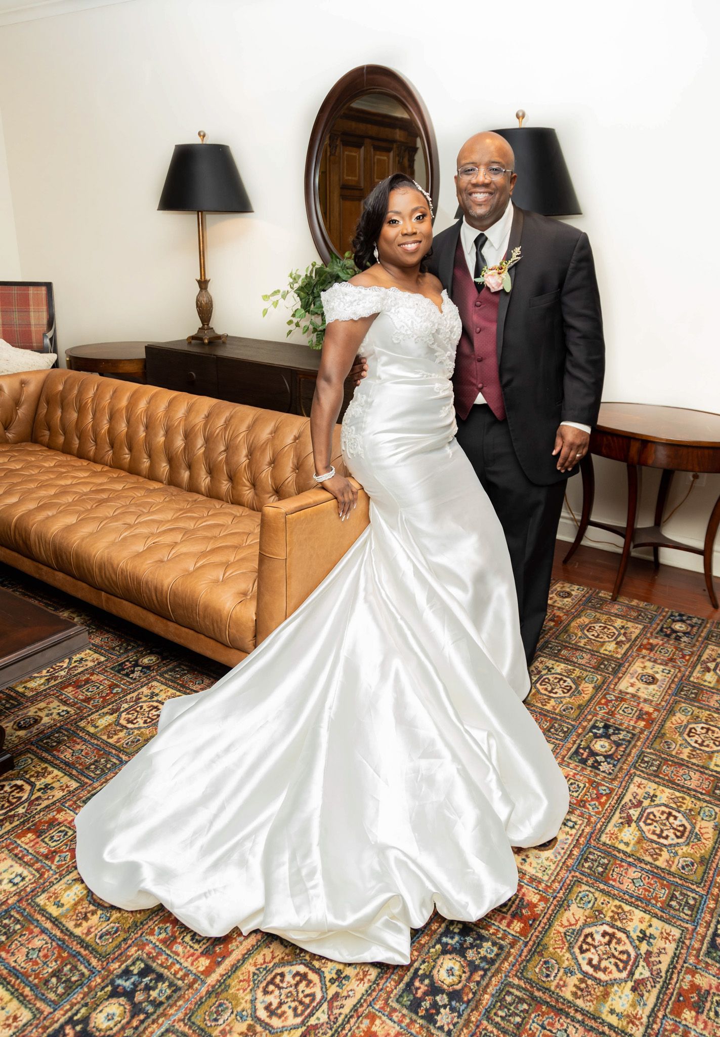 Elegant couple poses in formal wedding attire next to a brown leather sofa with classic decor.