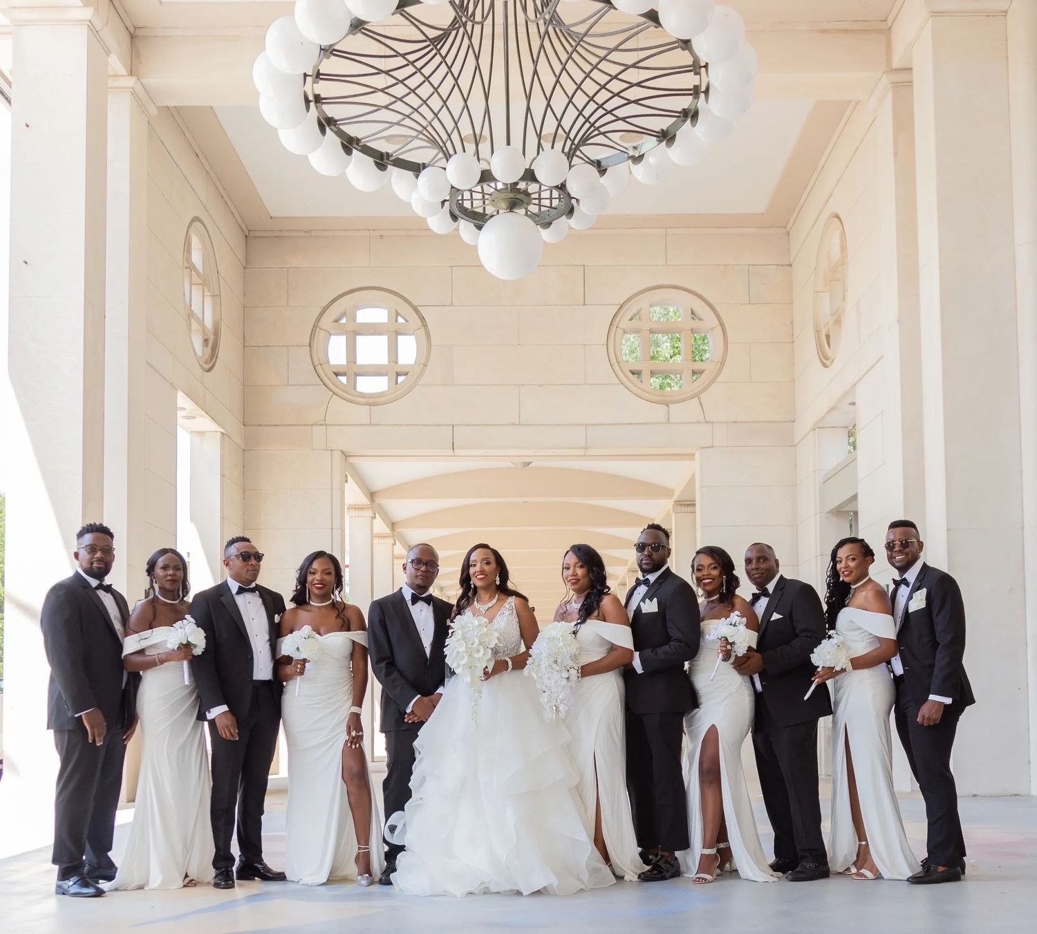 An elegant wedding party in black and white attire poses beneath a grand chandelier in a light-filled entrance hall.