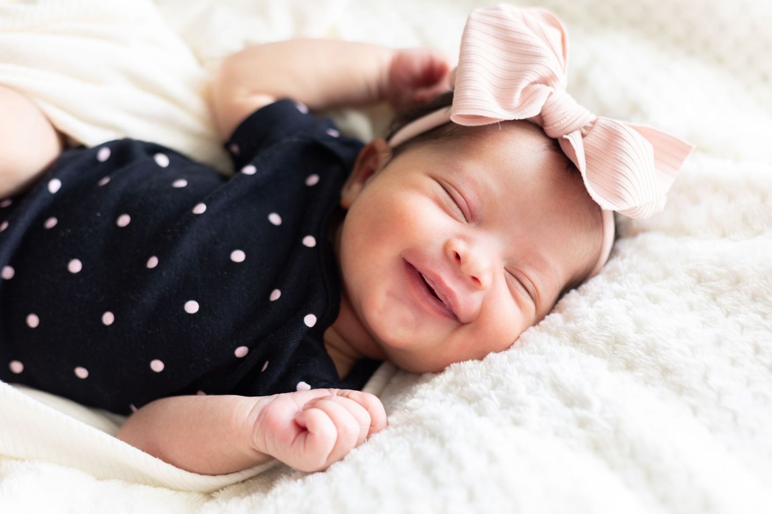 Sleeping newborn wearing navy polka dot outfit and pink bow on soft white blanket.