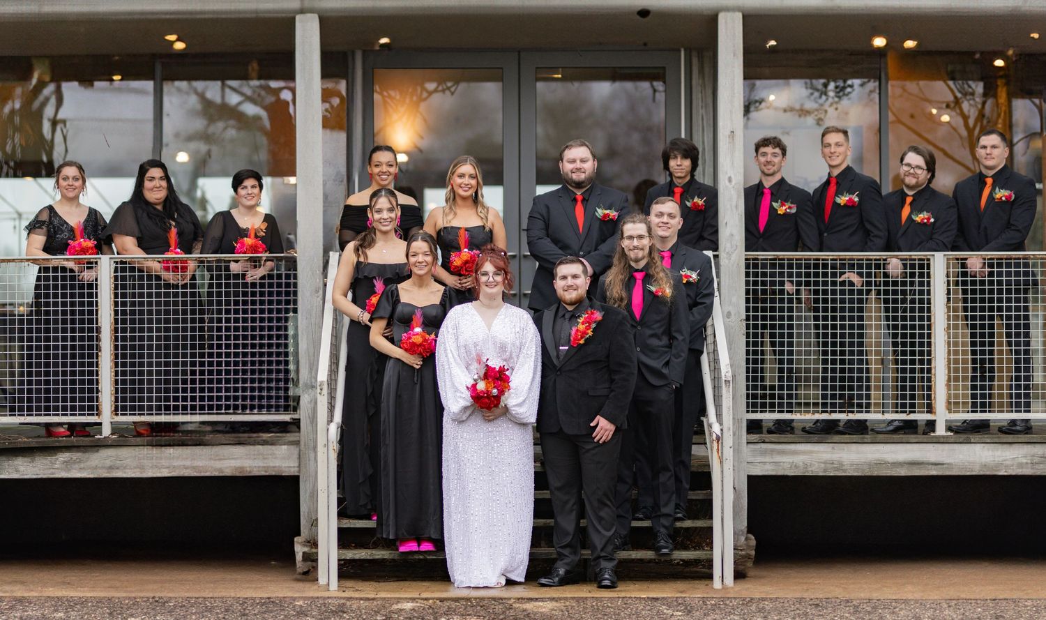 Wedding party poses on modern deck with bride in white dress surrounded by attendants in black.