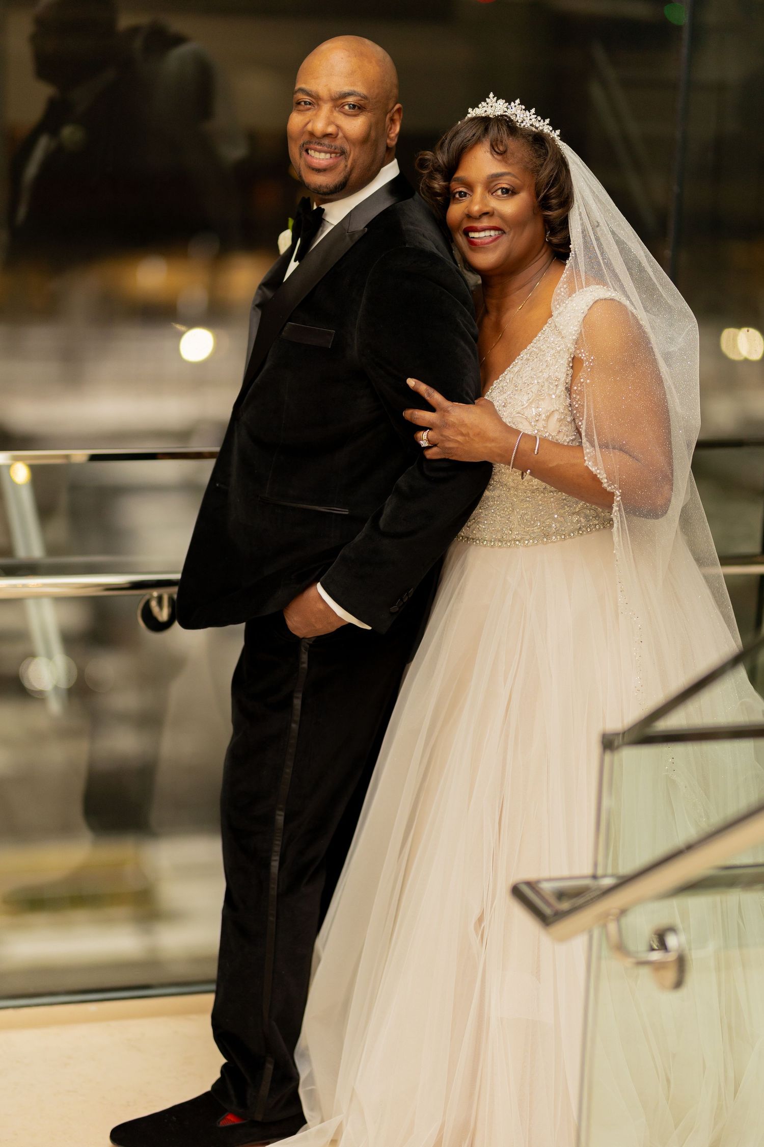 A couple stands on a staircase at their wedding, with the bride wearing a veil and tiara and the groom in a black tuxedo.