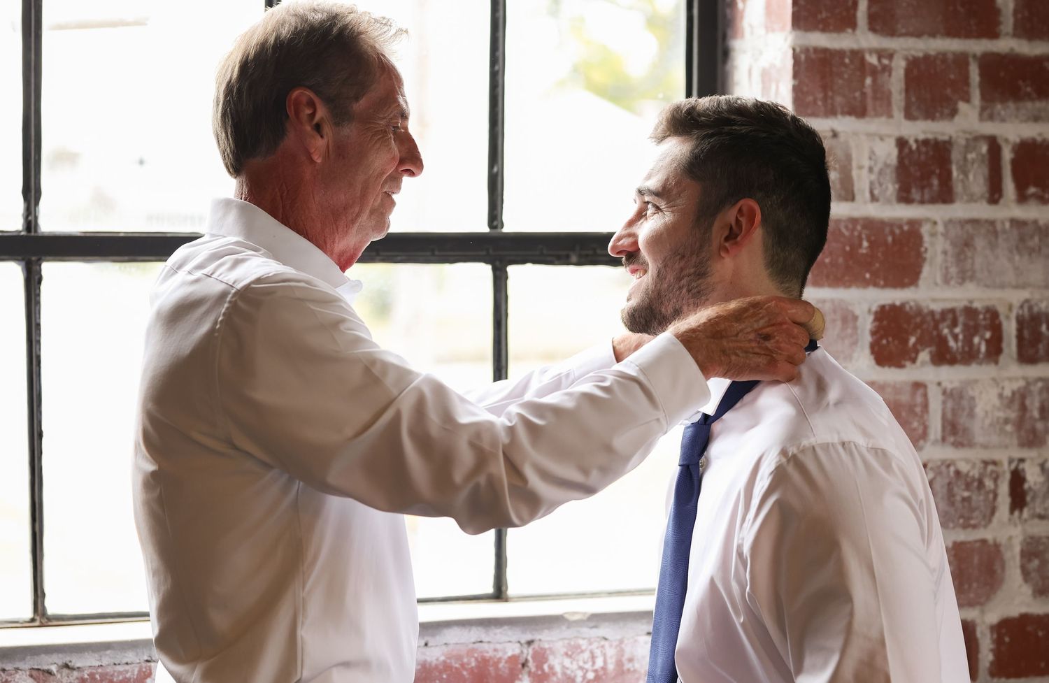Emotional moment between father and groom by window in brick venue.