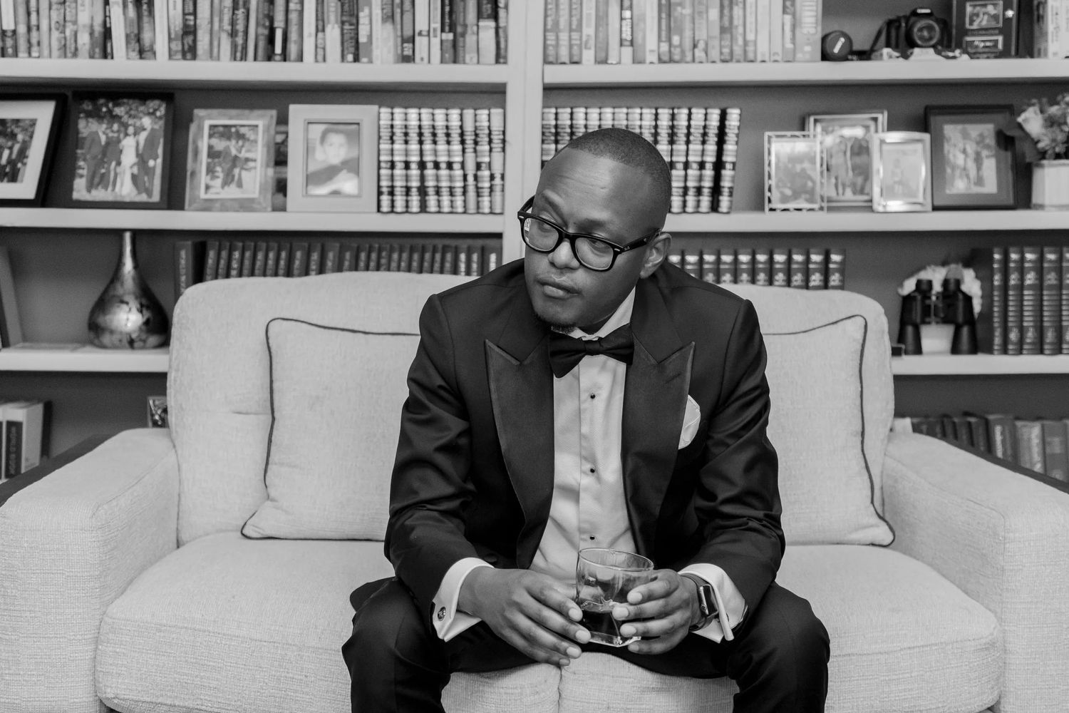 Man in glasses and suit sits thoughtfully on couch with bookshelf behind him in black and white photo.