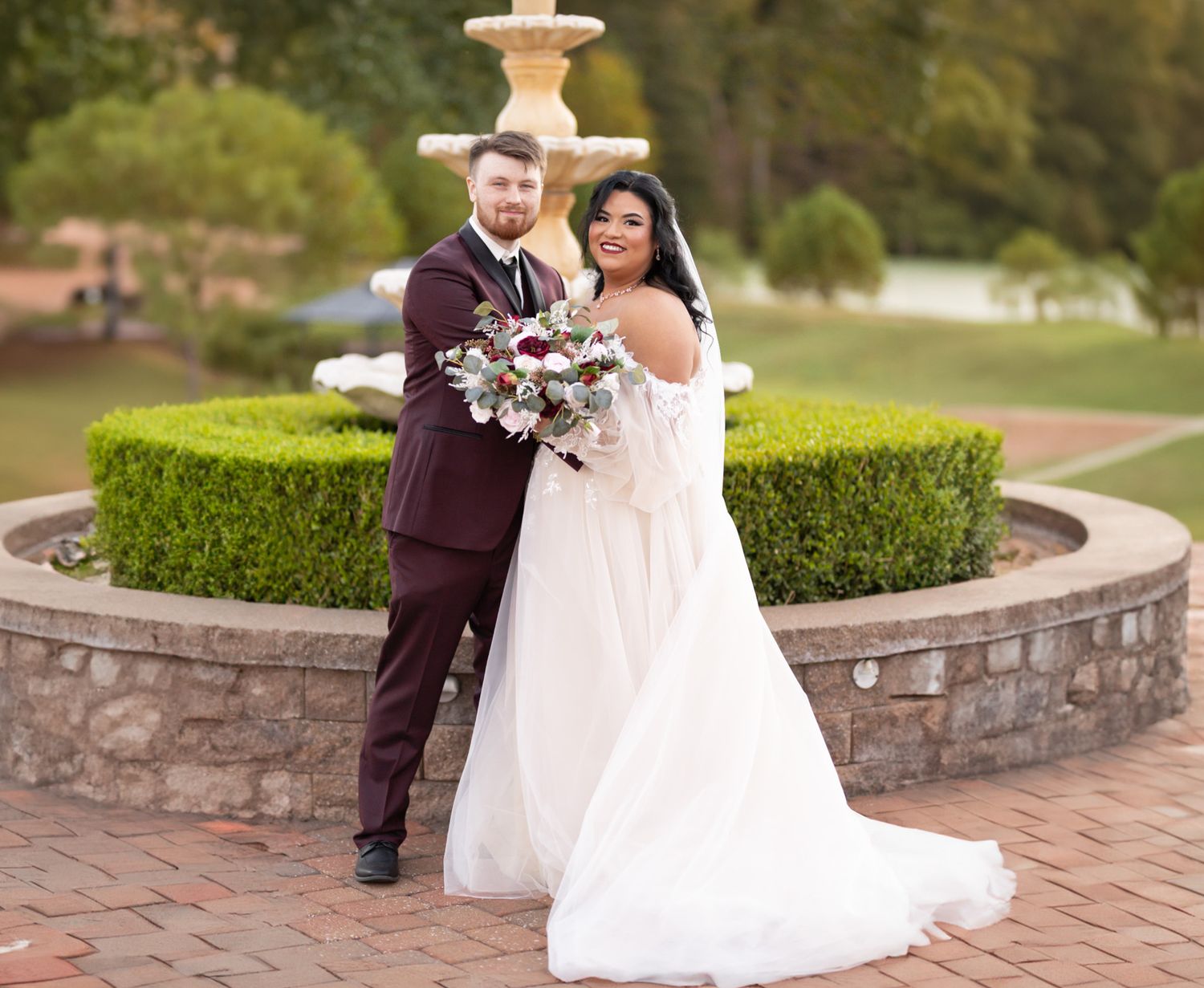 Newlyweds pose by an ornate stone fountain in a manicured garden.