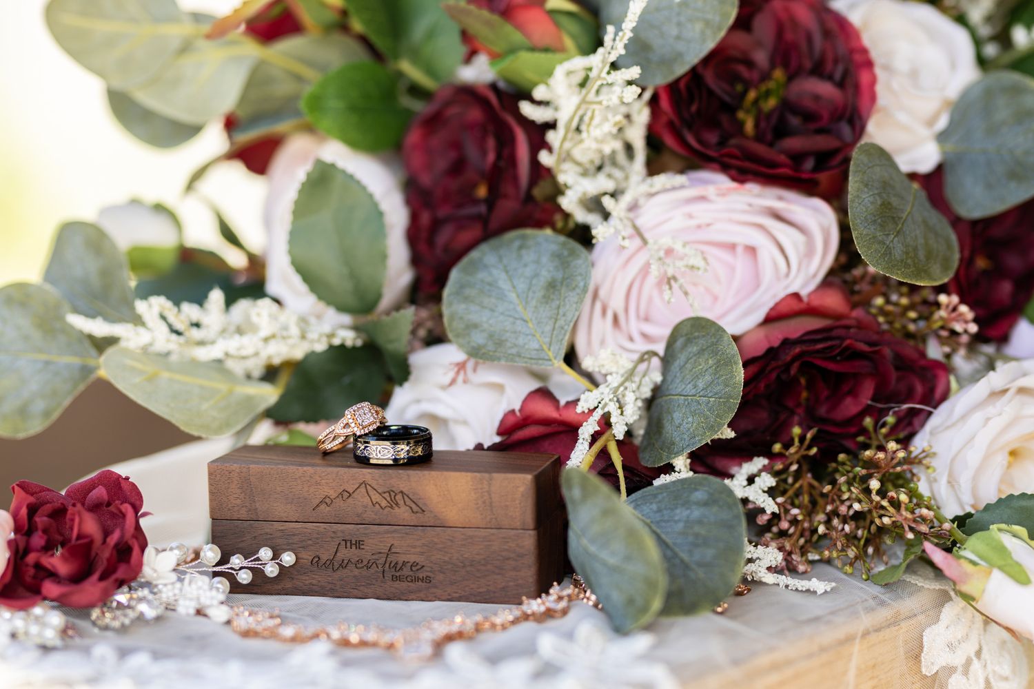 Wedding rings in wooden box surrounded by burgundy and pink roses with eucalyptus leaves on lace background.