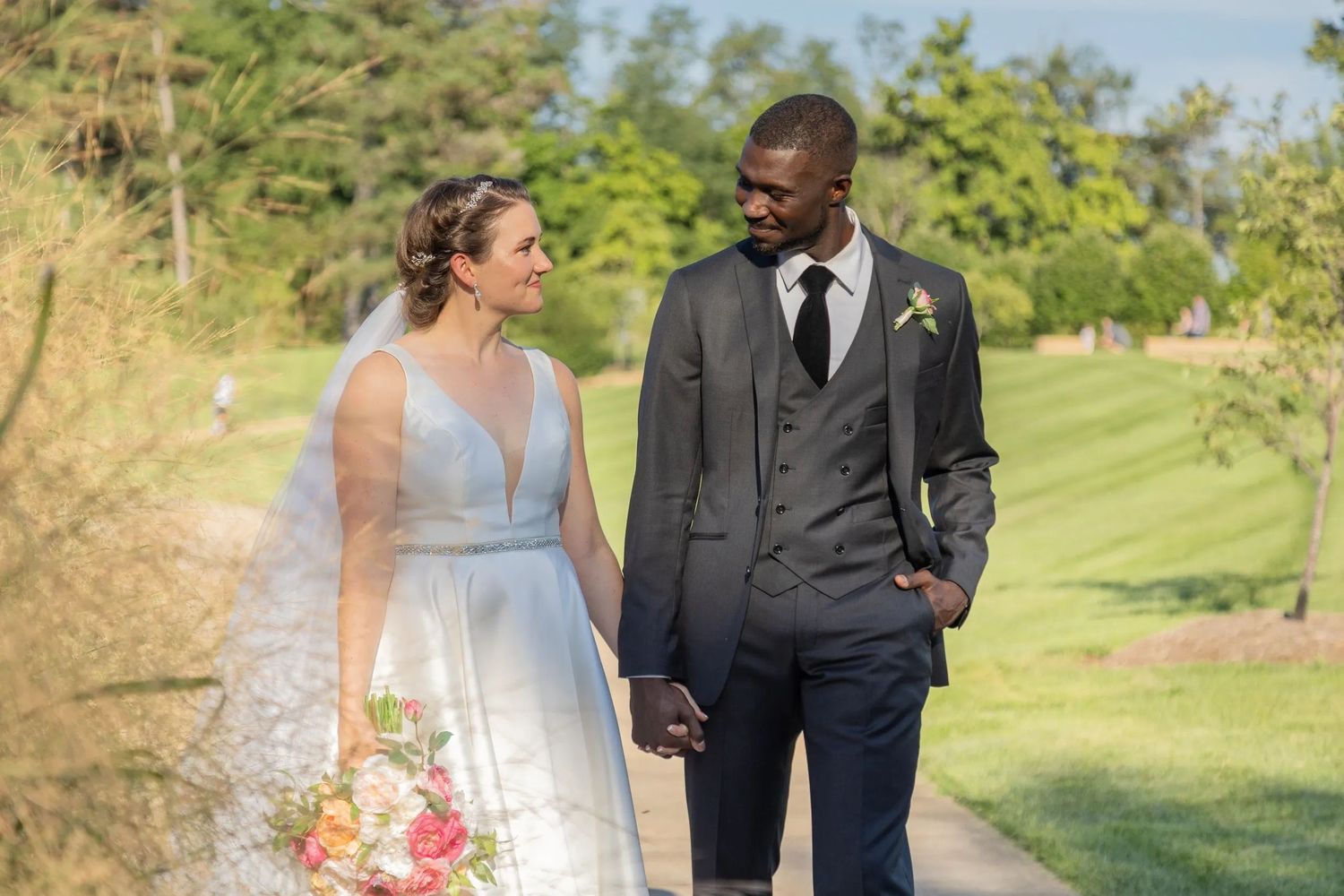 Couple in formal wedding attire walking and laughing together on a scenic garden path surrounded by lush greenery.
