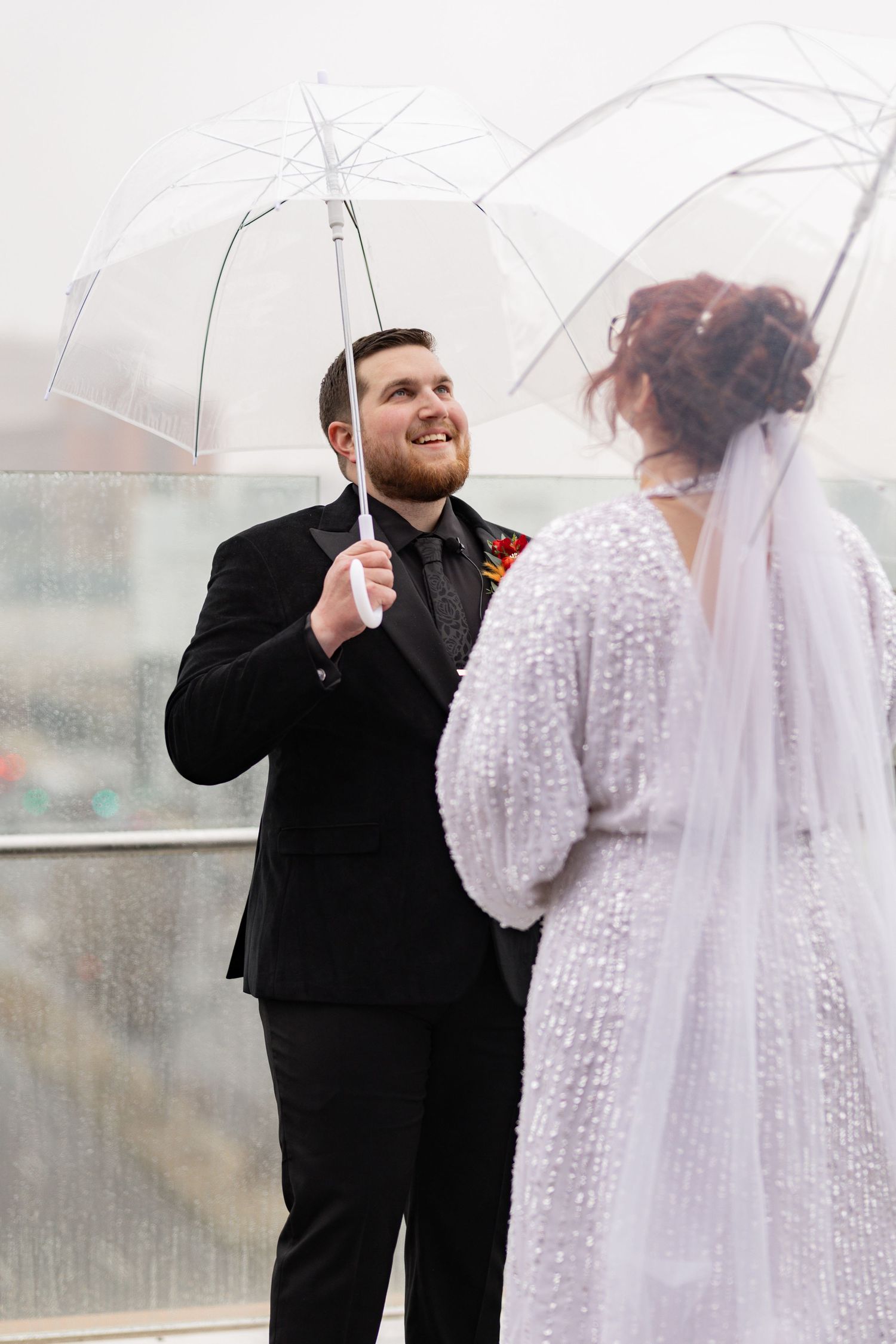 Couple shares a rainy wedding moment under clear umbrellas on a misty day.