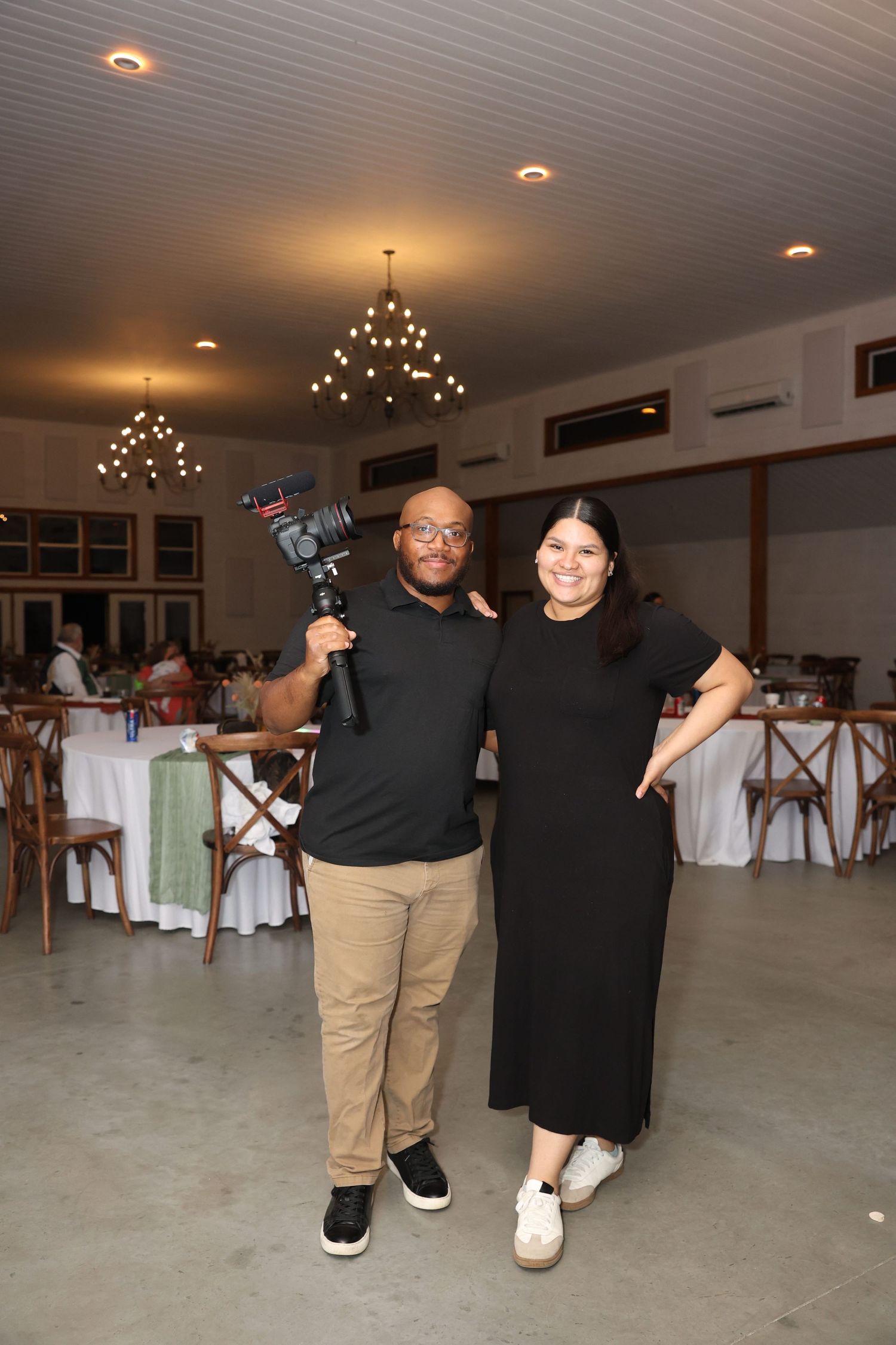 Two photographers in black attire stand together in an event venue with professional camera equipment.