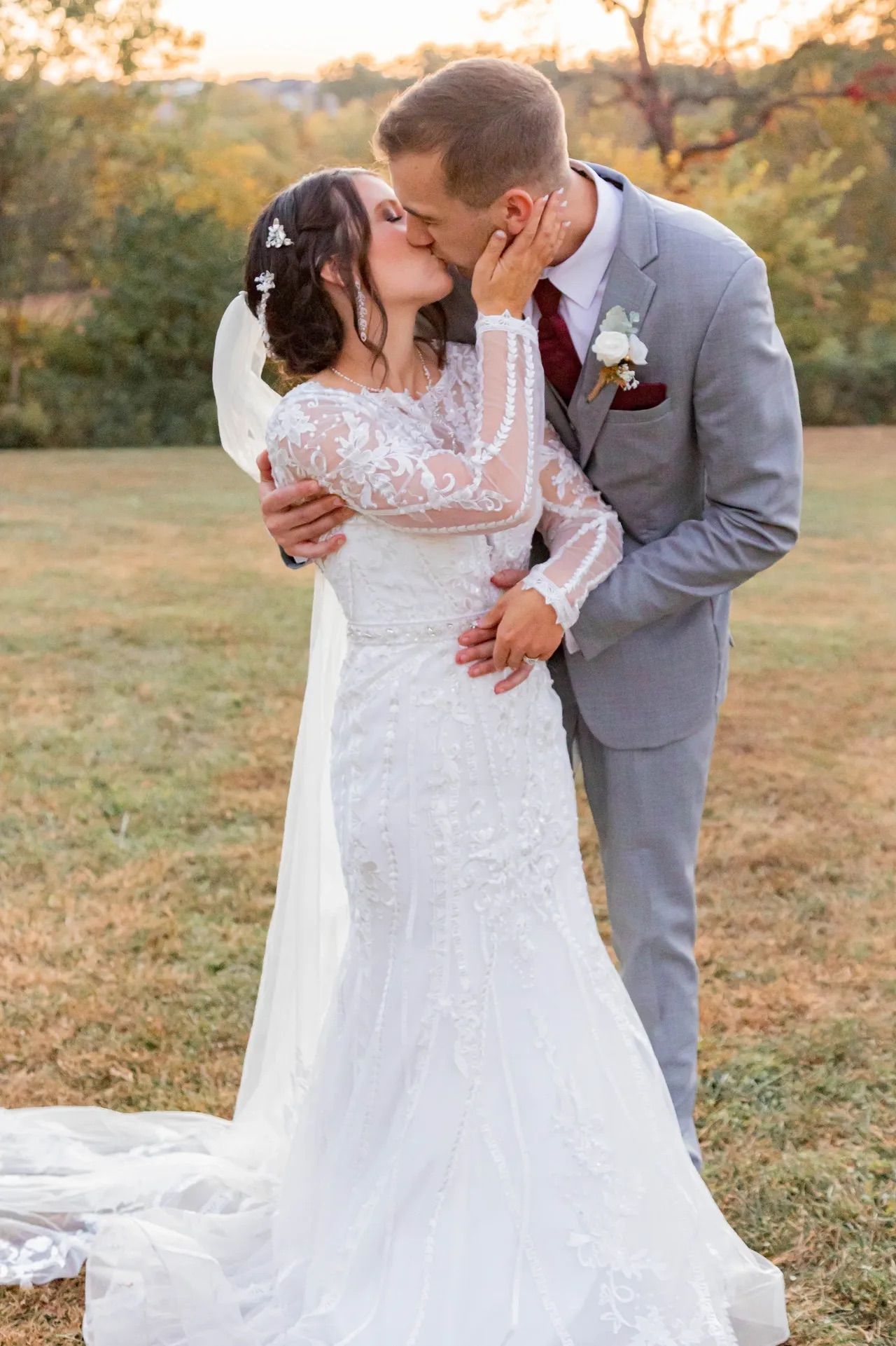 A romantic outdoor wedding scene at sunset with a bride in lace gown and groom in gray suit sharing a kiss.