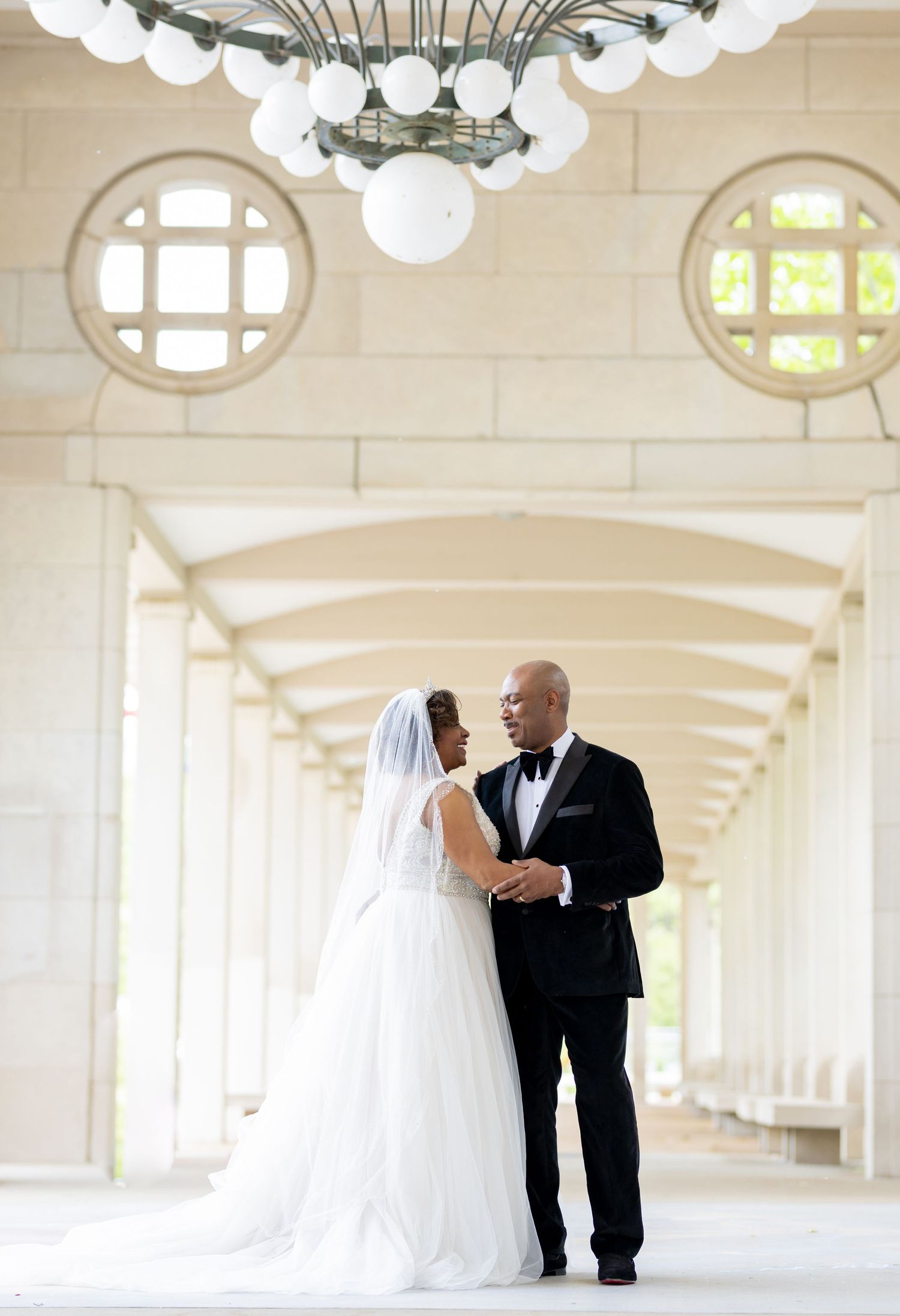 Elegant wedding photo of a couple in formal attire standing in a colonnade with dramatic lighting.