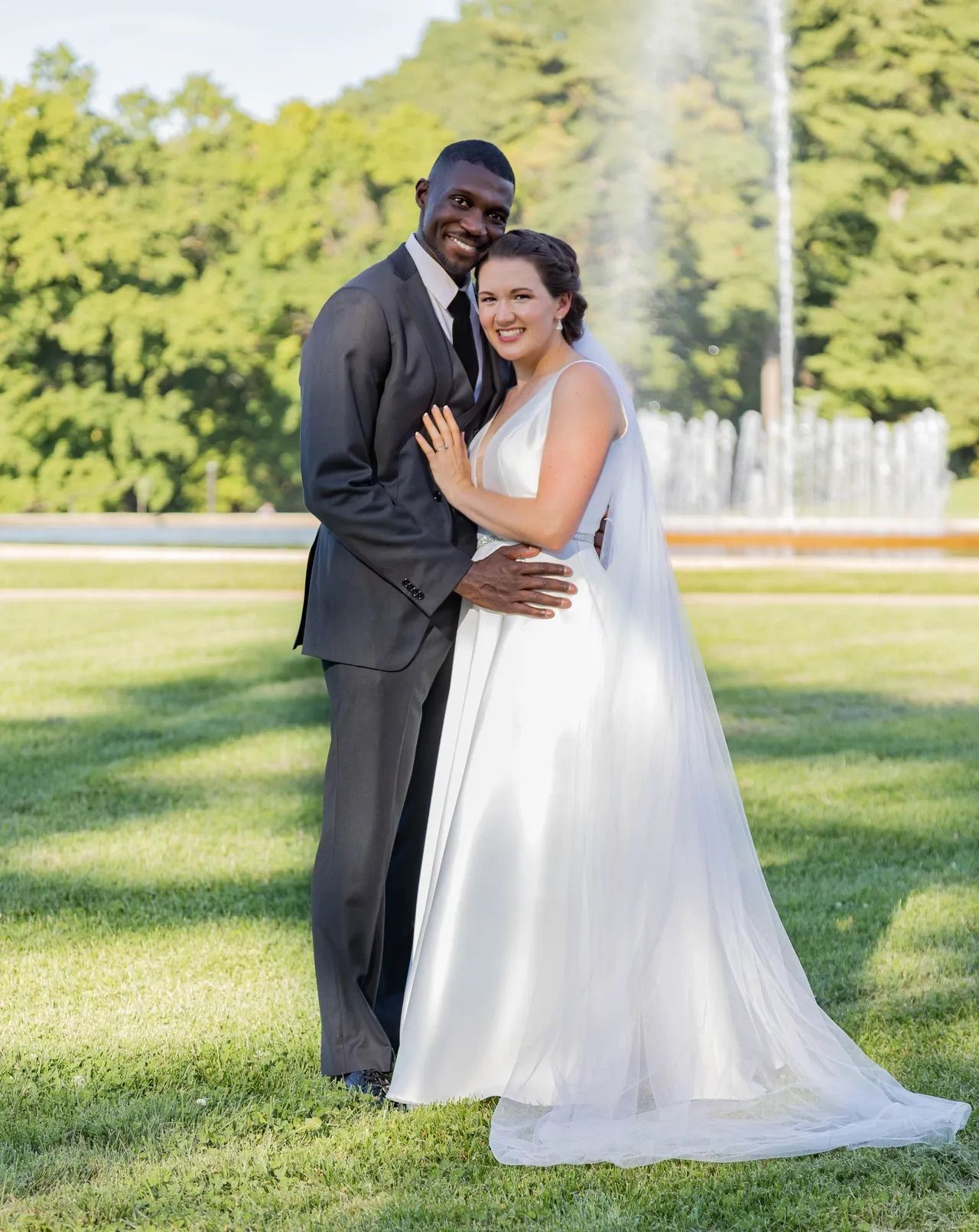 A groom and bride pose together outdoors on their wedding day with a fountain in the background.