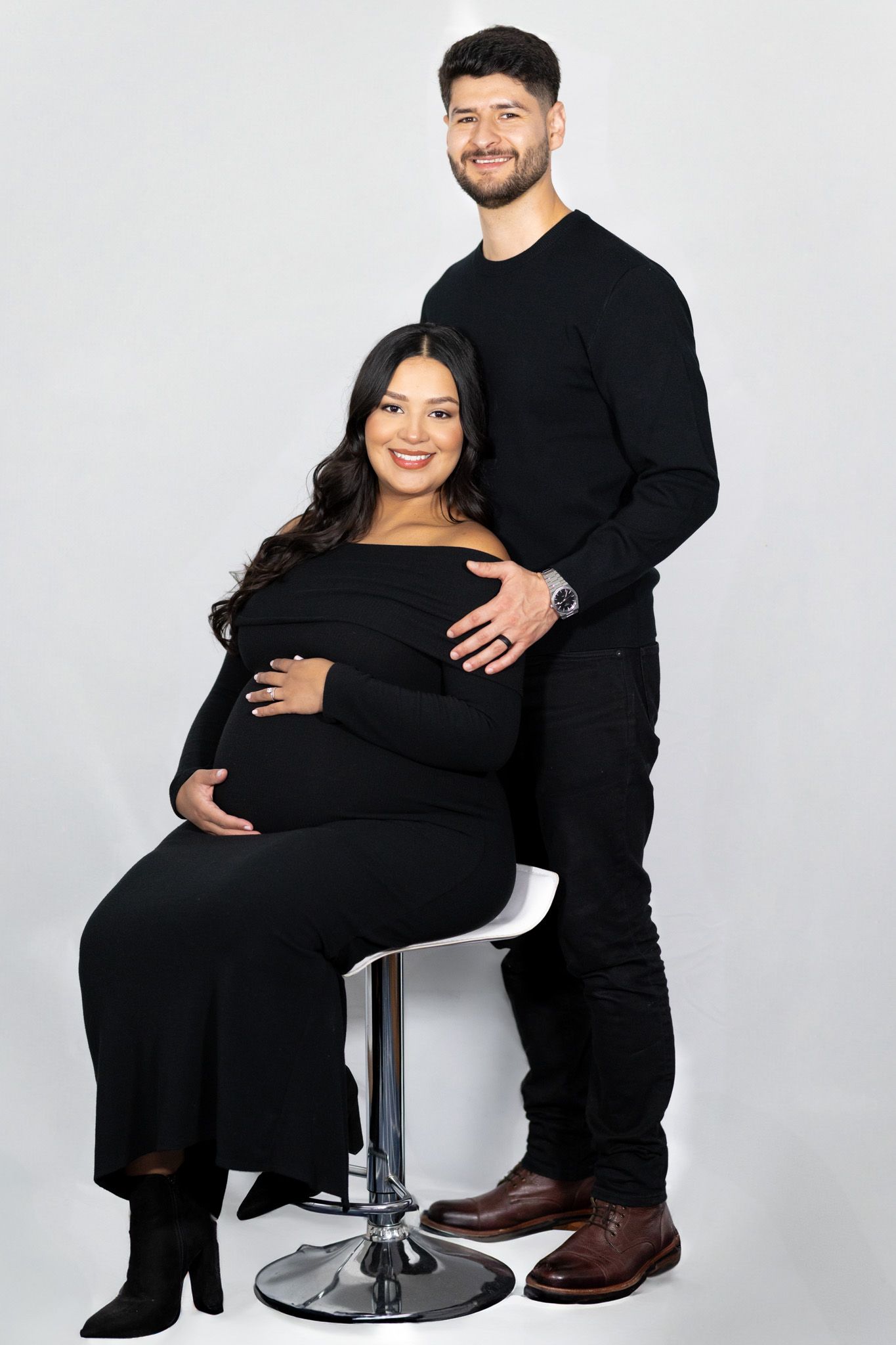 Expectant couple in matching black outfits posing on studio chair.