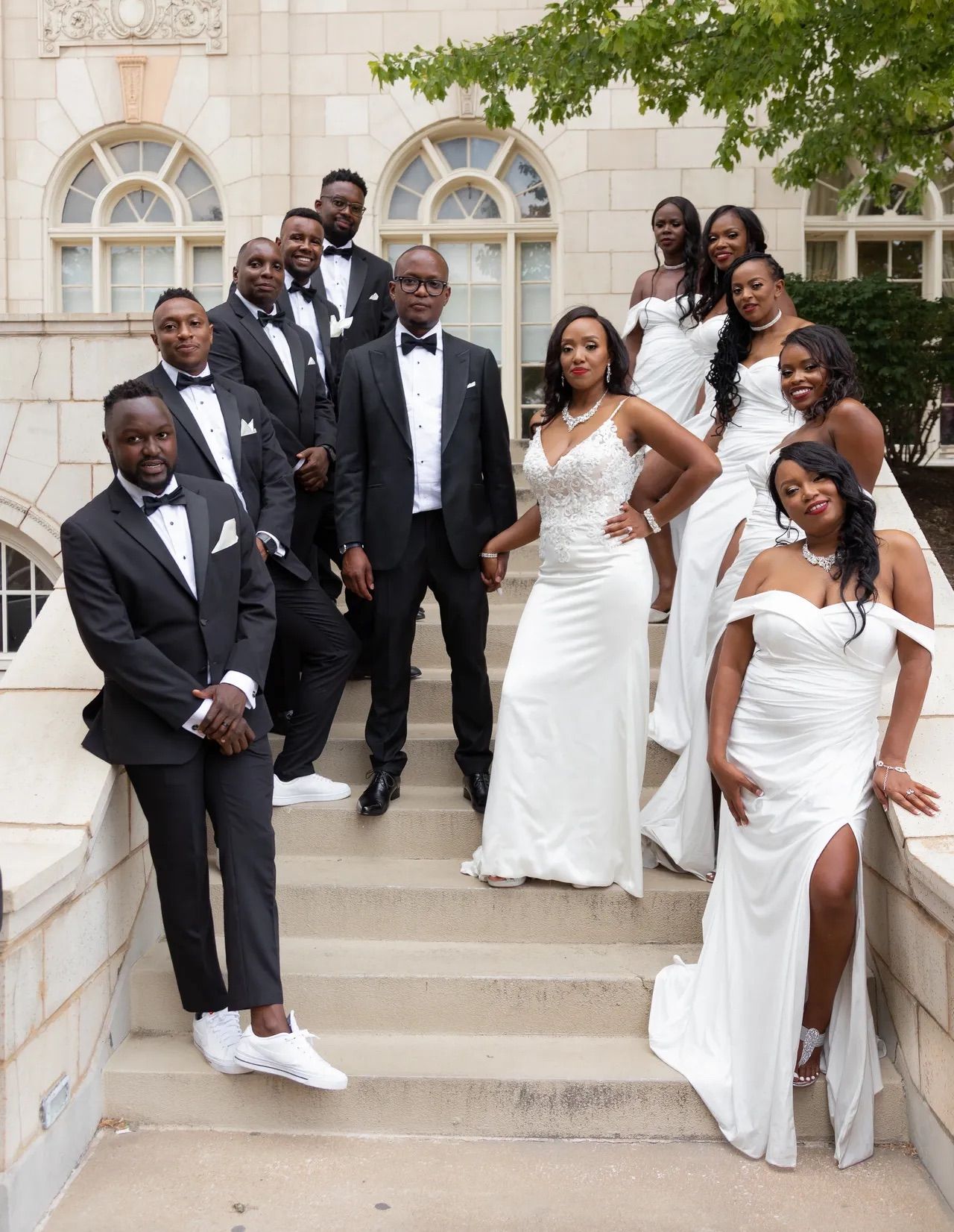 A wedding party posing in formal black and white attire on elegant stone steps outside a building with arched windows.