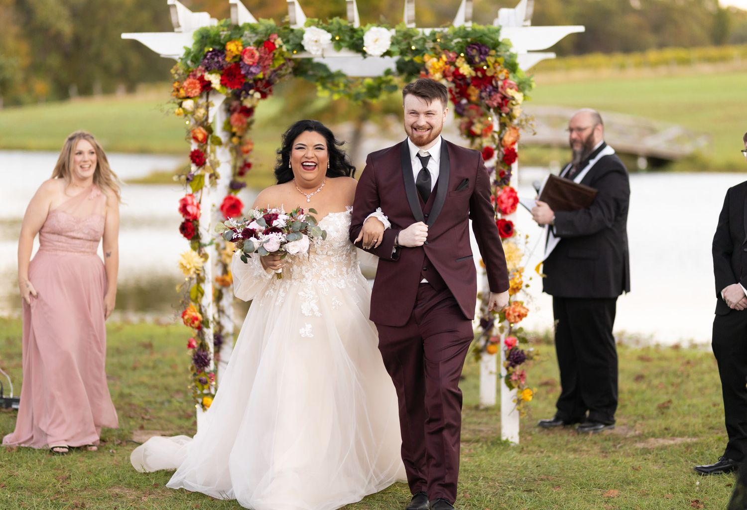 Wedding couple celebrates beneath a floral arch with burgundy roses and greenery at an outdoor ceremony.