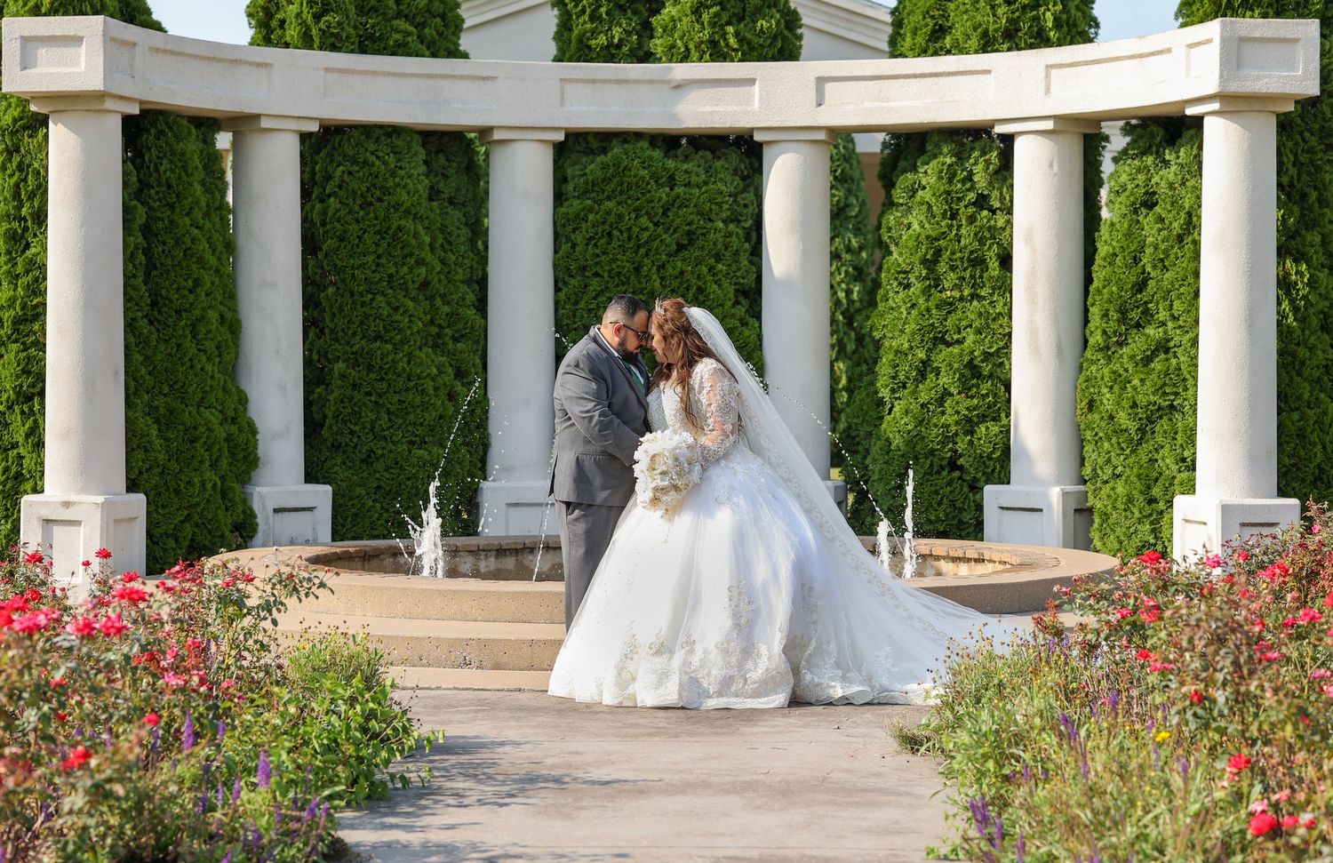 A romantic wedding moment under an elegant white pergola surrounded by blooming red flowers and green hedges.