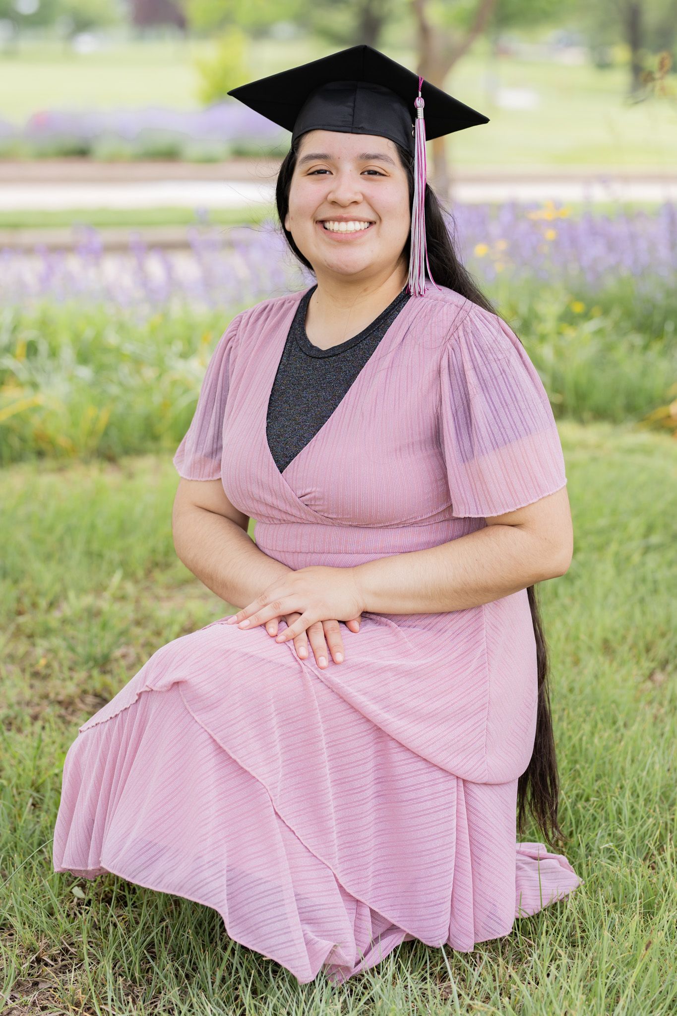 A graduation portrait in a garden setting with purple flowers featuring a pink dress and graduation cap.