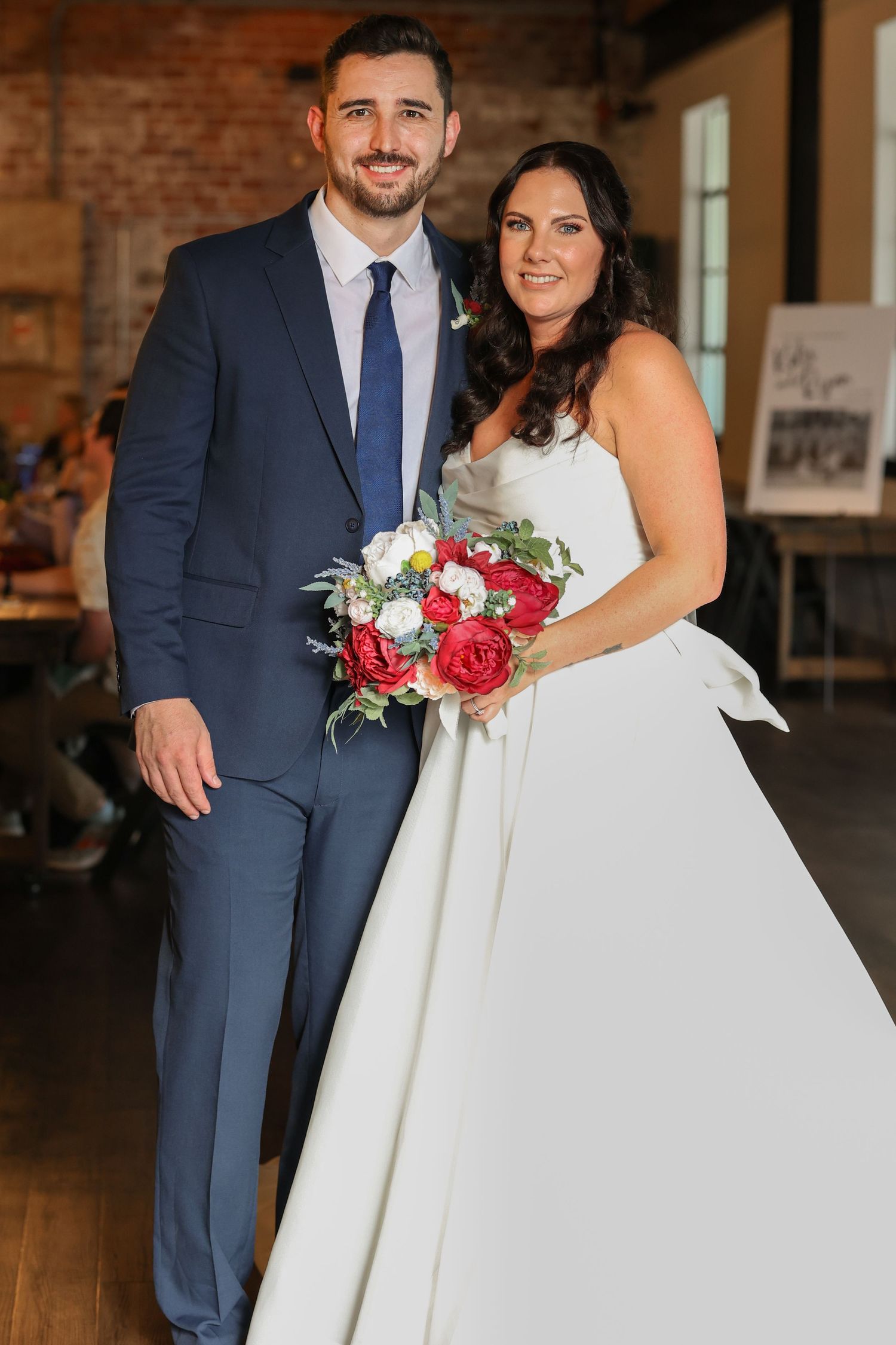 Wedding couple poses in rustic brick venue with red and white bouquet.