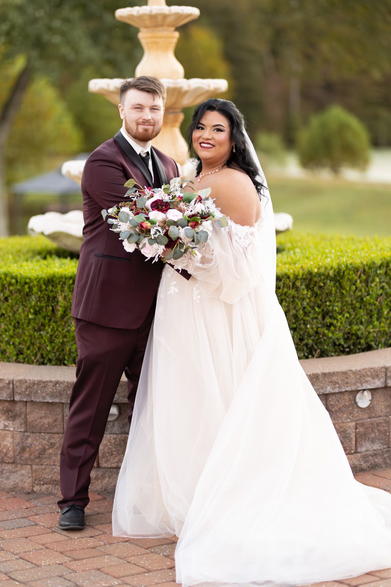 A romantic wedding couple poses together outdoors on a brick patio surrounded by manicured hedges and greenery.