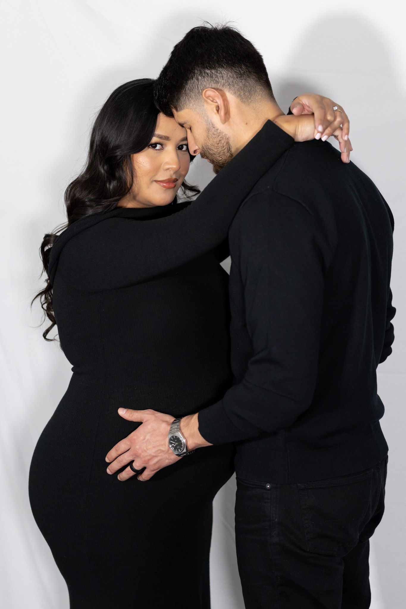 Intimate maternity portrait of couple in black clothing against white backdrop.