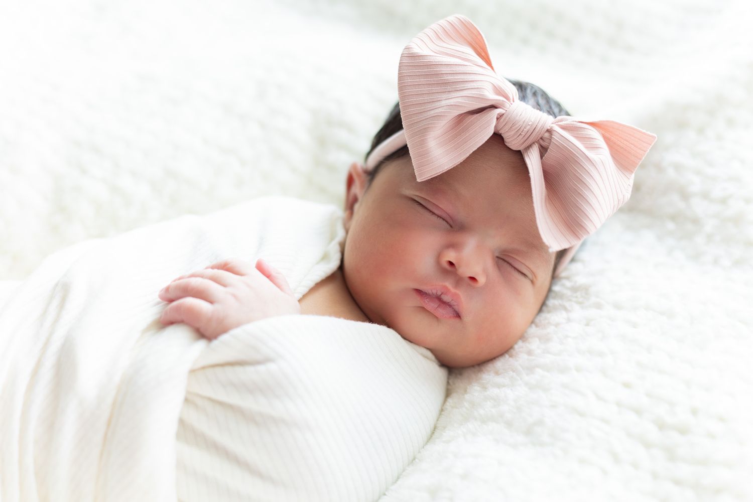 Peaceful sleeping baby wearing pink bow headband on white blanket.