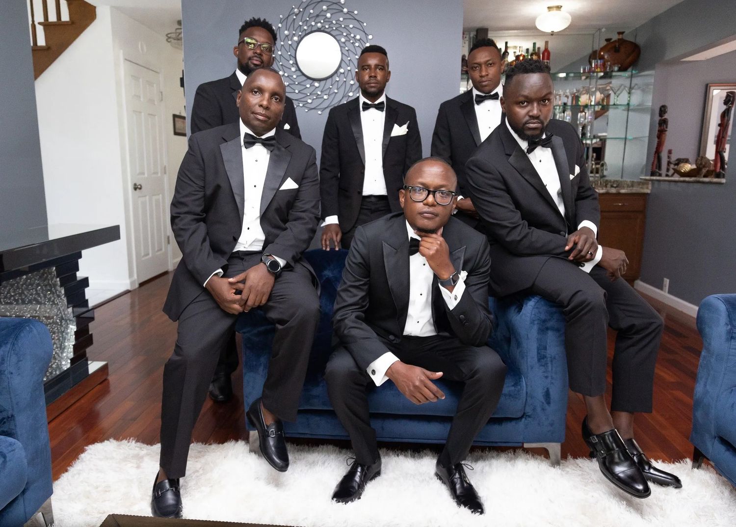 A group in formal black tuxedos and bow ties pose together on a blue couch in an elegant home interior.