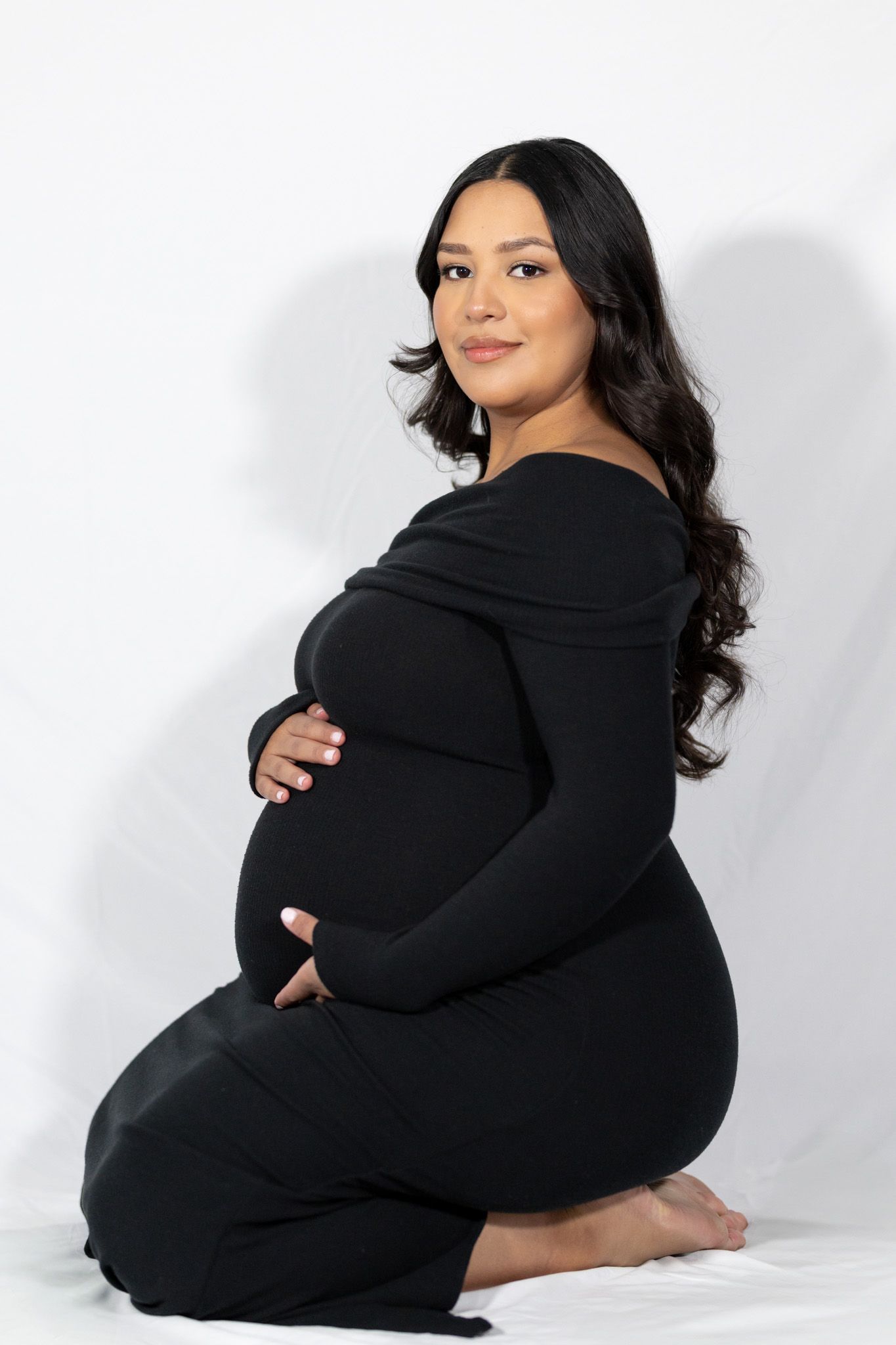 Side profile maternity portrait in black off-shoulder dress against white background.
