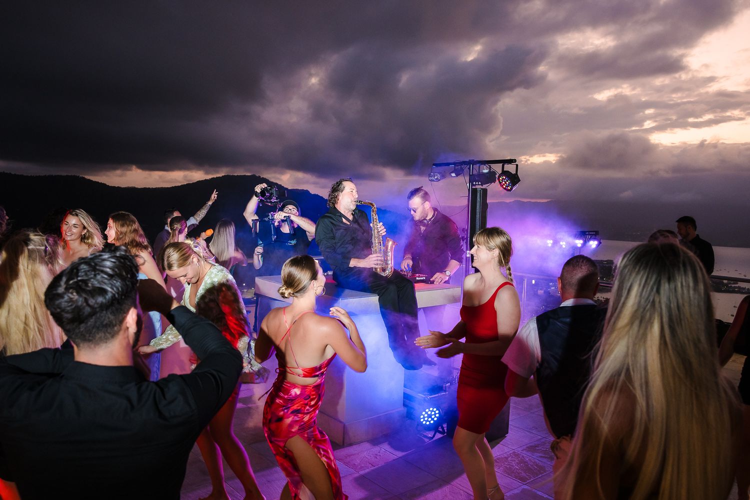 People dancing and celebrating at a wedding reception with dramatic stormy clouds and purple lighting effects in the background.