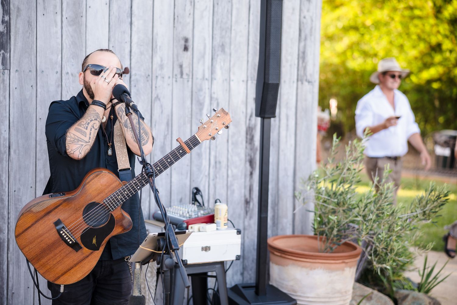 A musician plays acoustic guitar against a gray wall while singing into a microphone outdoors.