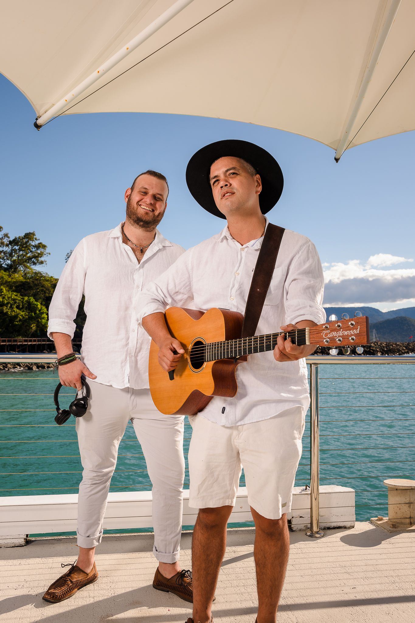 Two musicians in white outfits perform on a dock by turquoise water, one playing acoustic guitar under a white umbrella.