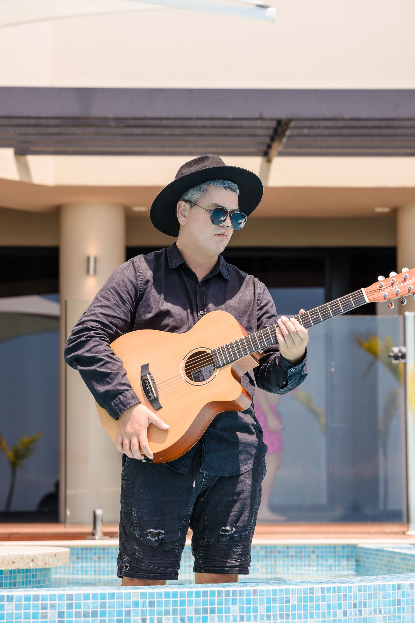 Musician in black attire and sunglasses plays acoustic guitar while standing by a swimming pool.