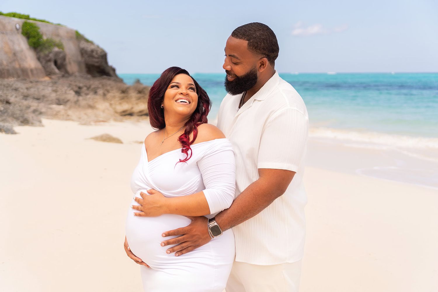 Maternity photo of couple in white clothing embracing on tropical beach.