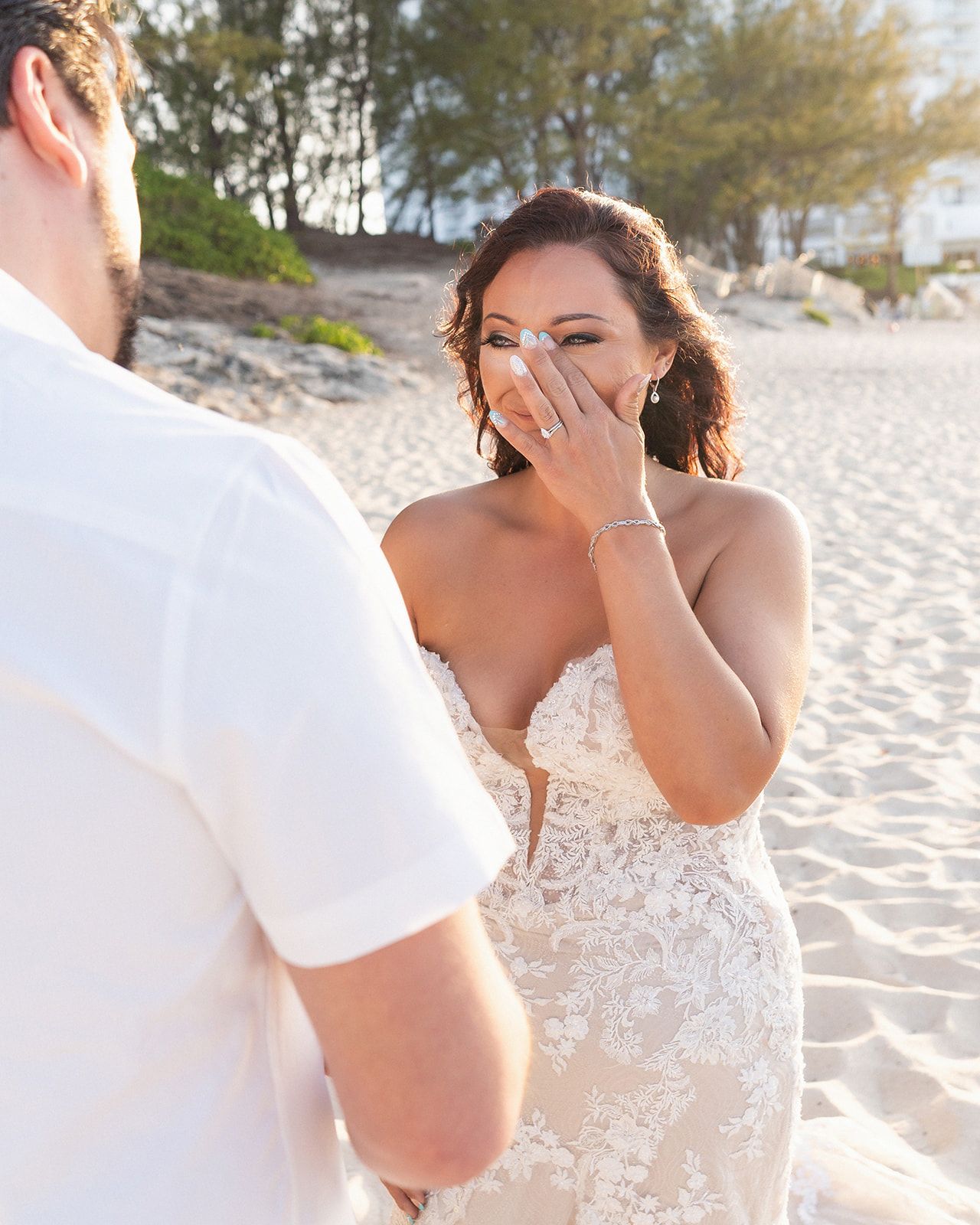 A joyful emotional reaction is captured during a beach photoshoot as someone in an elegant lace wedding gown covers their mouth.