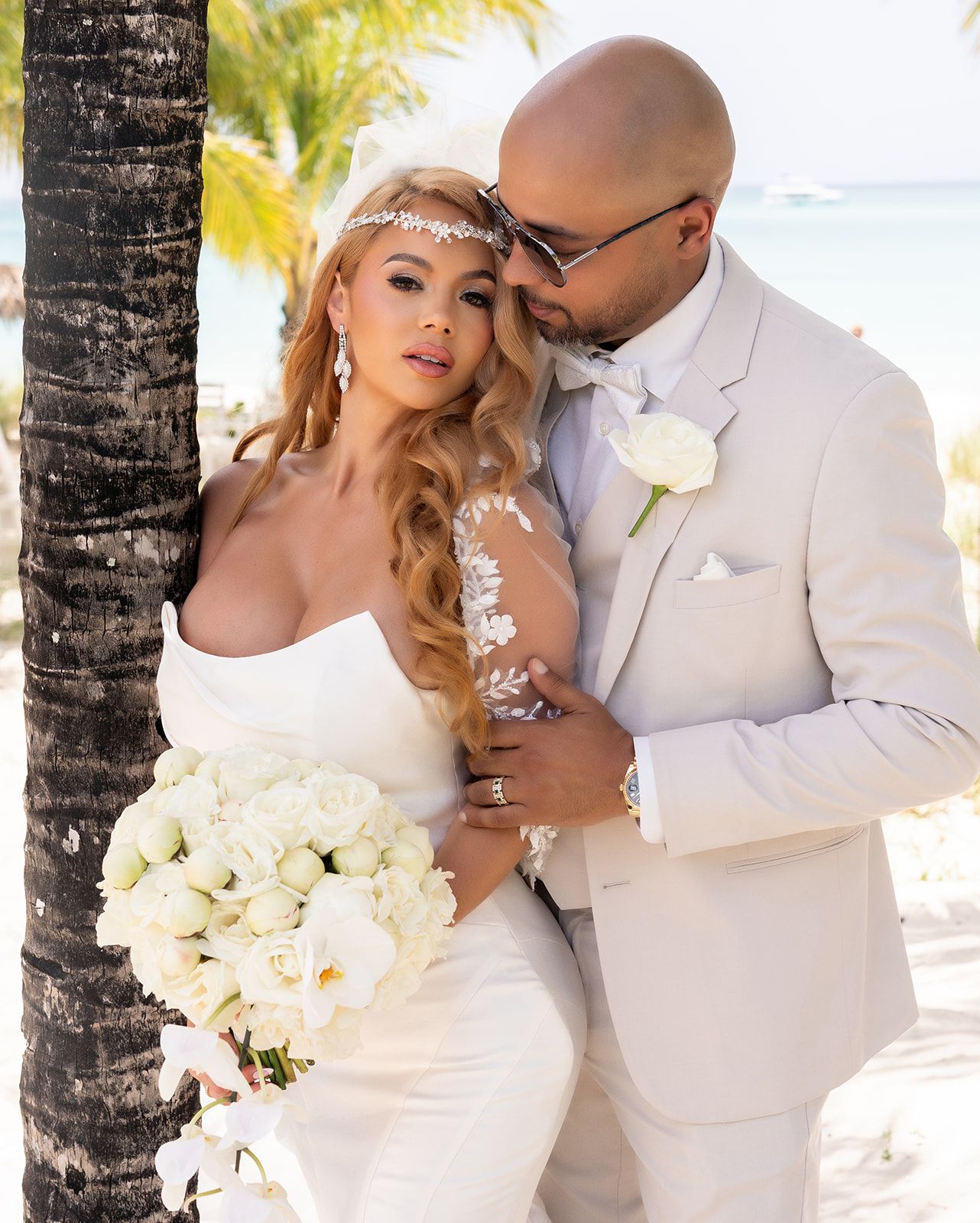A romantic beach wedding couple embraces by a palm tree, with the bride holding a white rose bouquet and wearing lace.