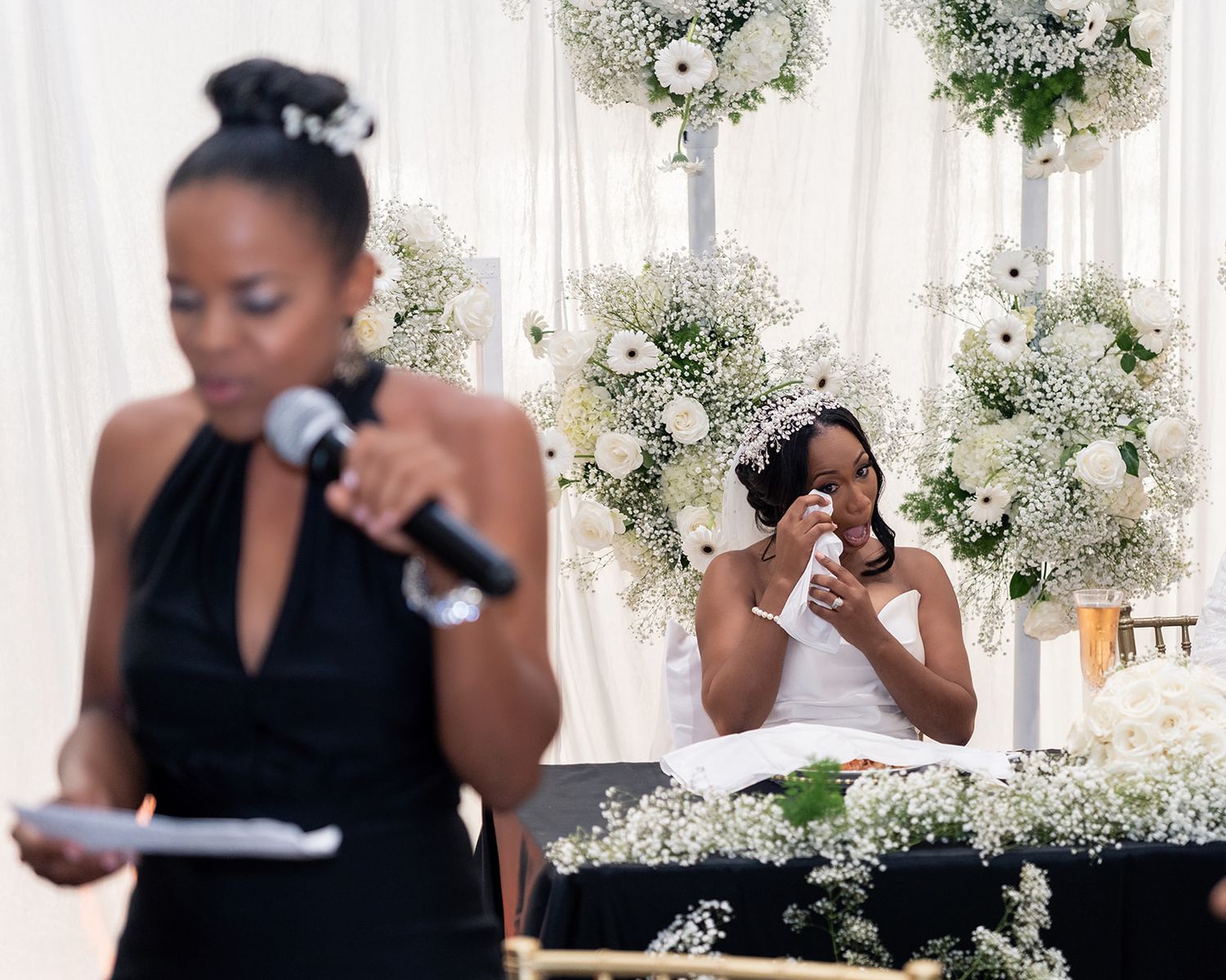 Two people giving emotional speeches at a wedding reception decorated with white floral arrangements.