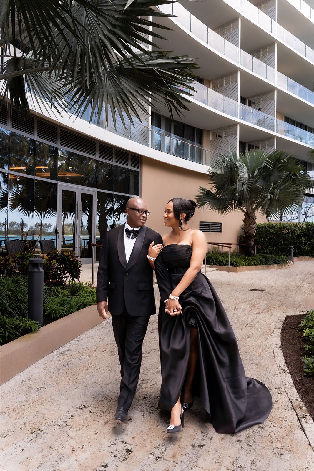A couple in formal black attire poses outside a modern beachfront hotel with palm trees.