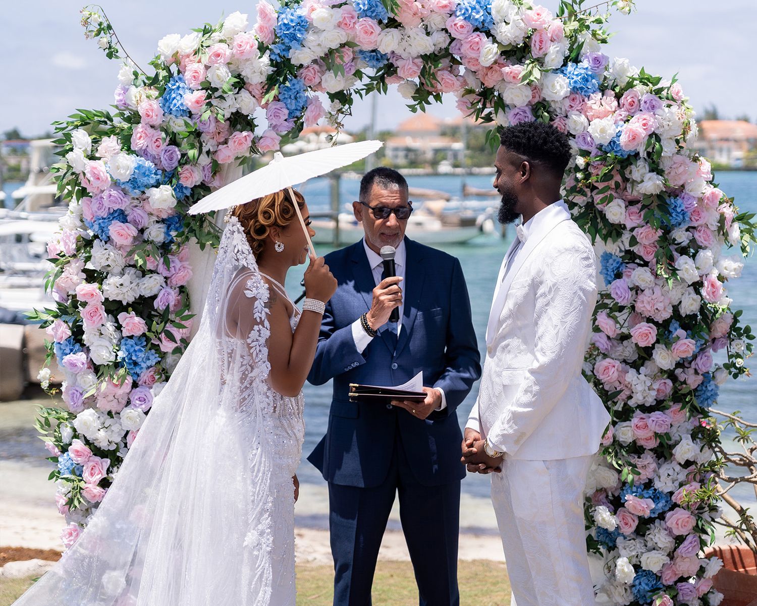 Wedding ceremony under a beautiful floral arch with pink and blue flowers by the waterfront during a sunny day.