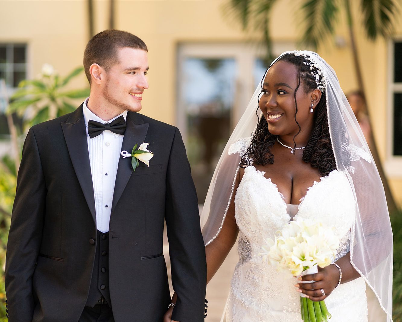 Newlywed couple wearing black tuxedo and white wedding dress smiles at each other during outdoor photo session.