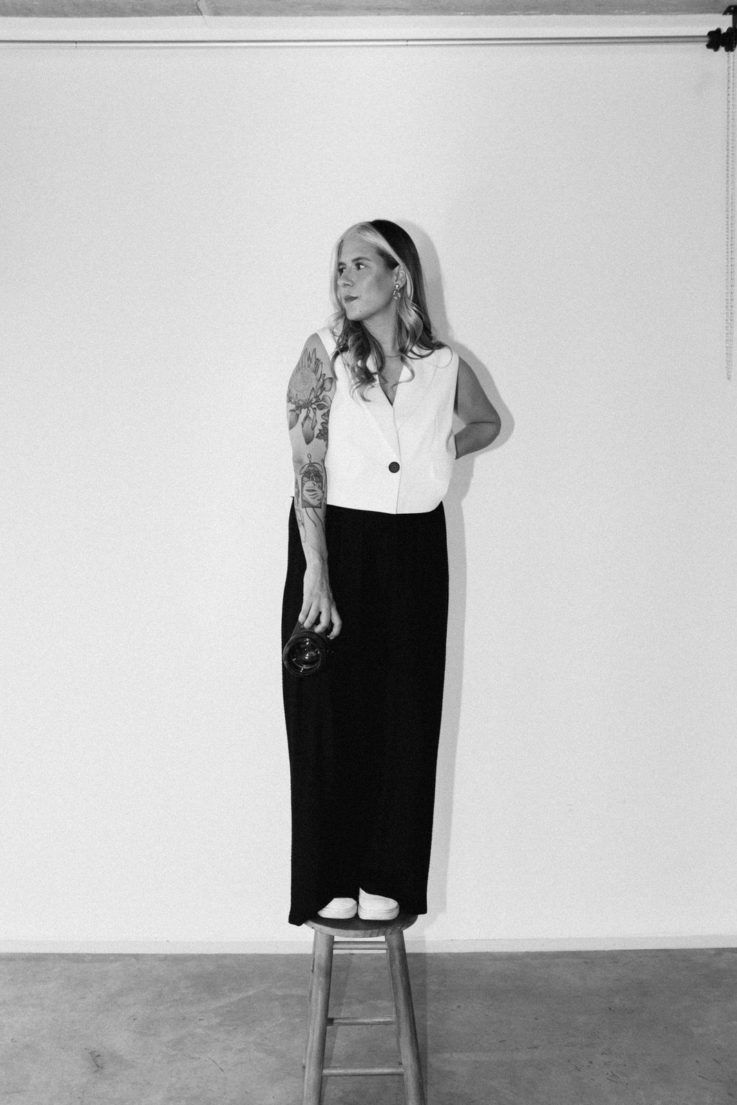 A person in minimalist black and white clothing stands on a stool in a studio setting.