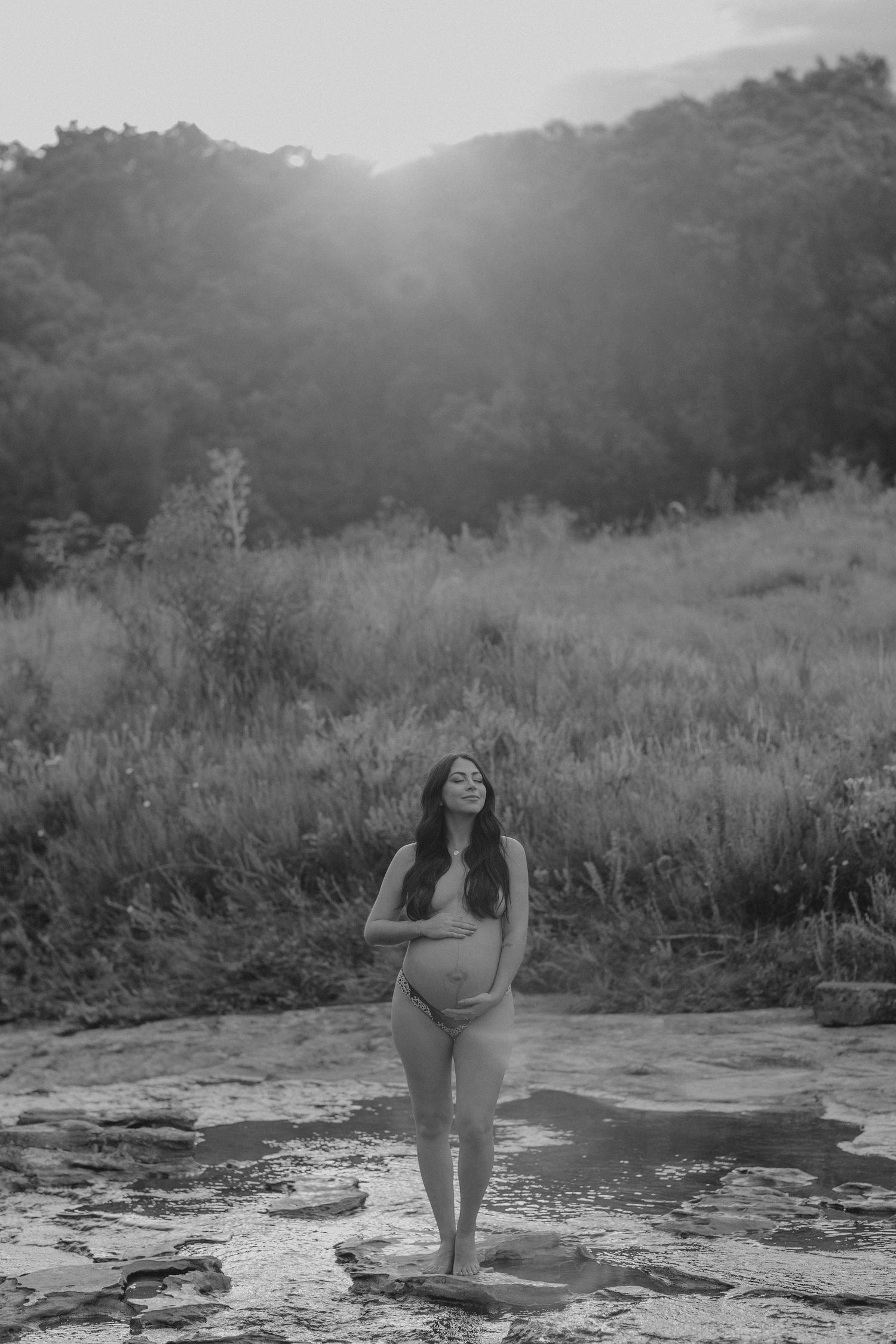 A figure stands at sunset on rocky terrain with mountains and wild grass in the background in black and white.