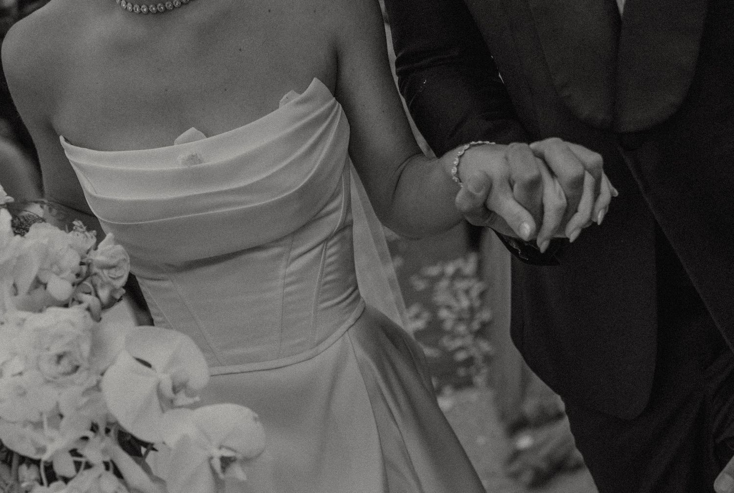 Black and white photo showing a bride in a white strapless dress holding hands with someone in formal wear.
