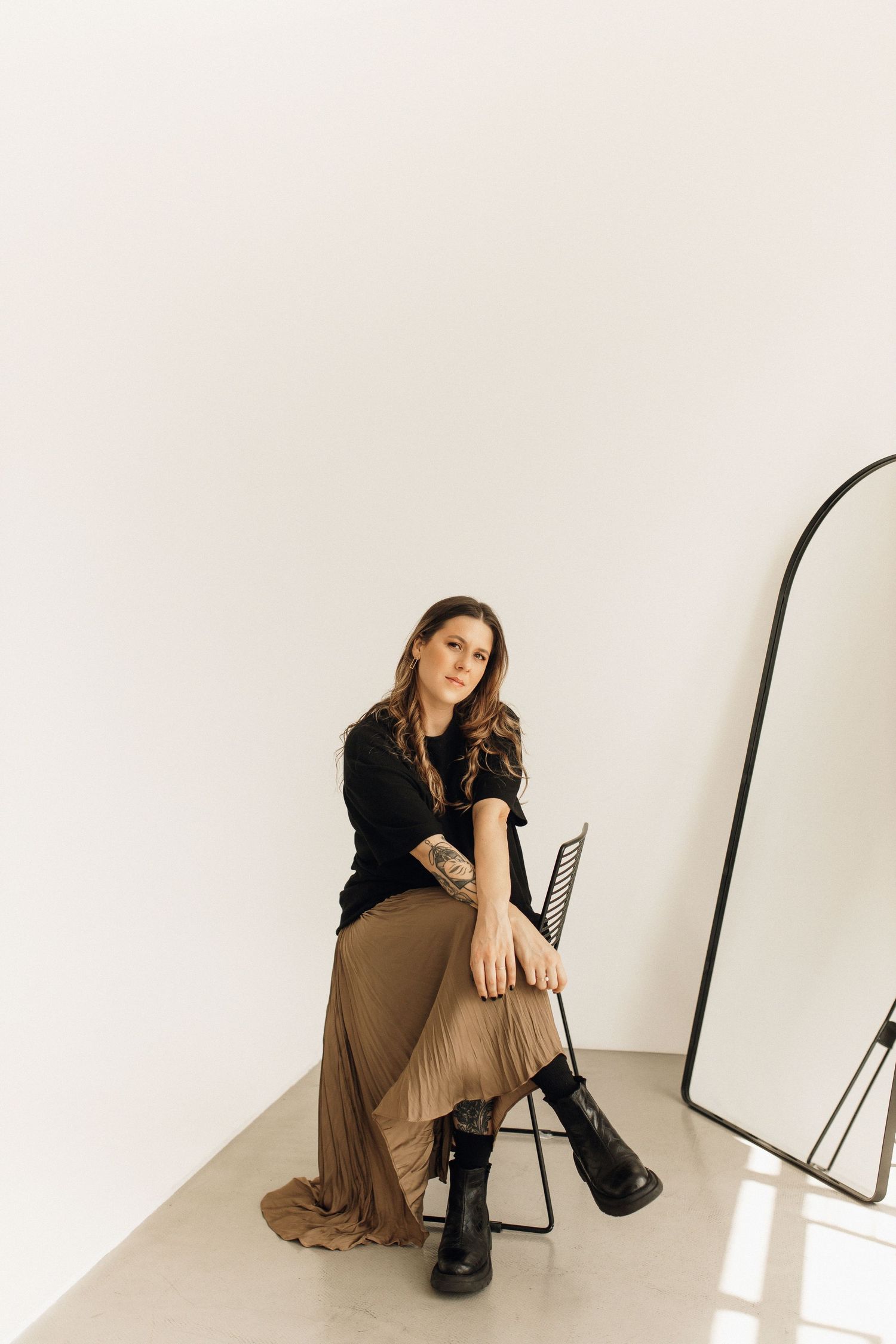 A person seated on a chair poses in a long skirt and black top near a modern mirror.