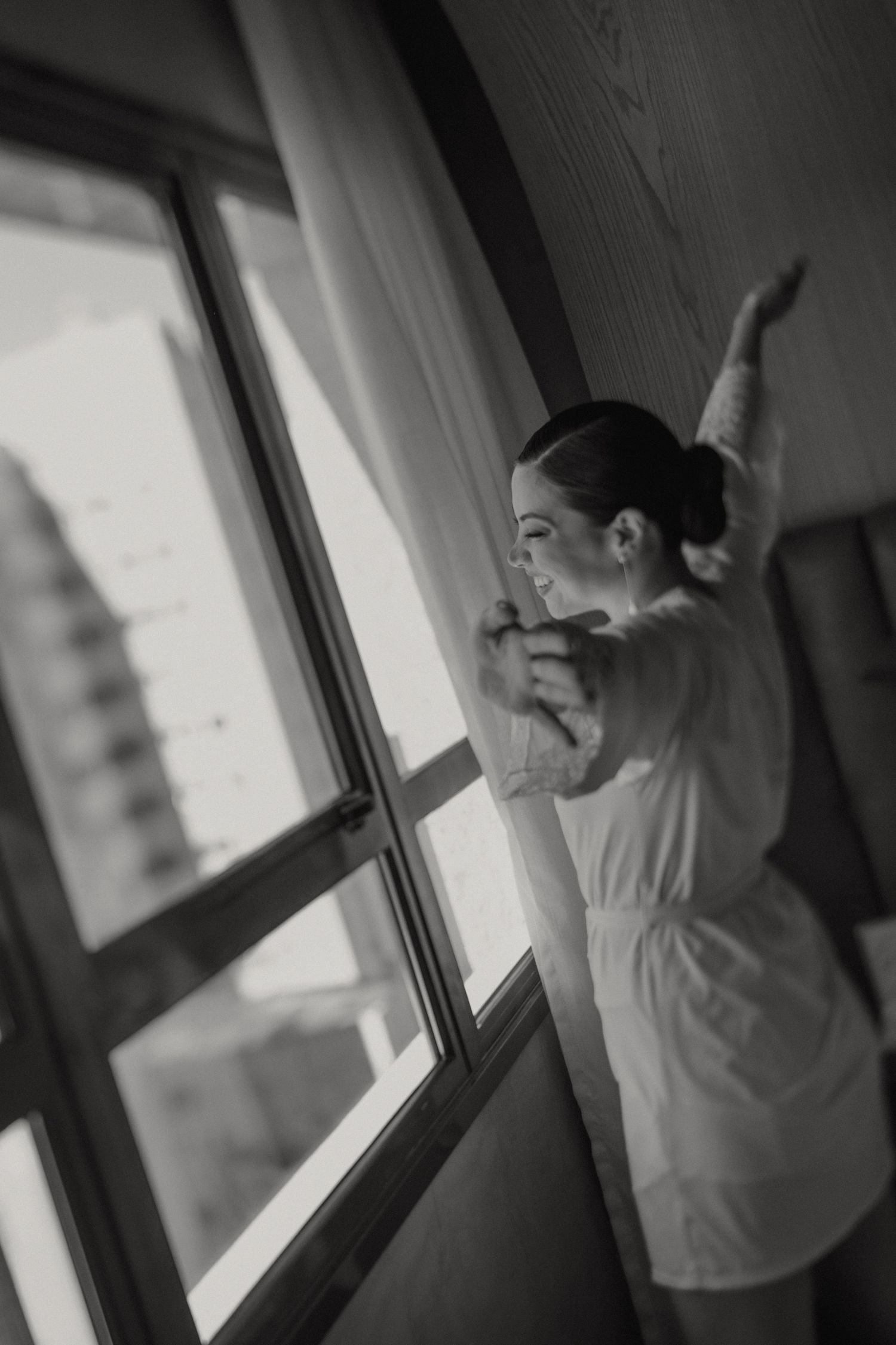 A grayscale photograph of a ballet dancer gazing through a window while wearing a flowing white dress.