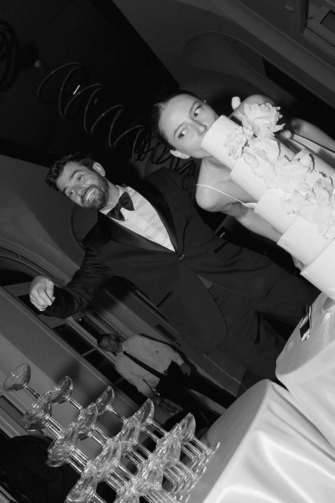 Black and white candid photo of a bride and groom sharing a playful moment during their wedding reception near champagne glasses.