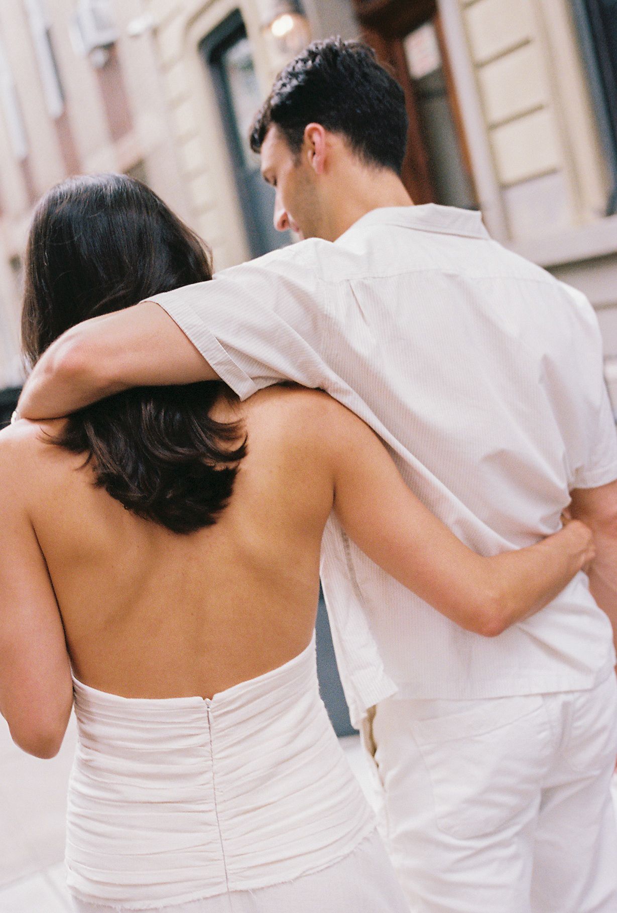 Back view of couple in white attire walking arm in arm down a street.