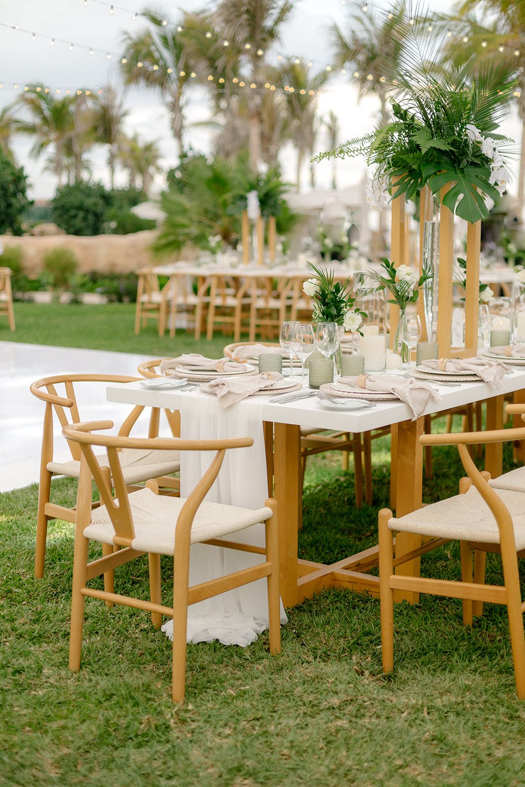 Elegant outdoor wedding reception setup with wooden Wishbone chairs and tropical palm centerpieces on rustic tables.
