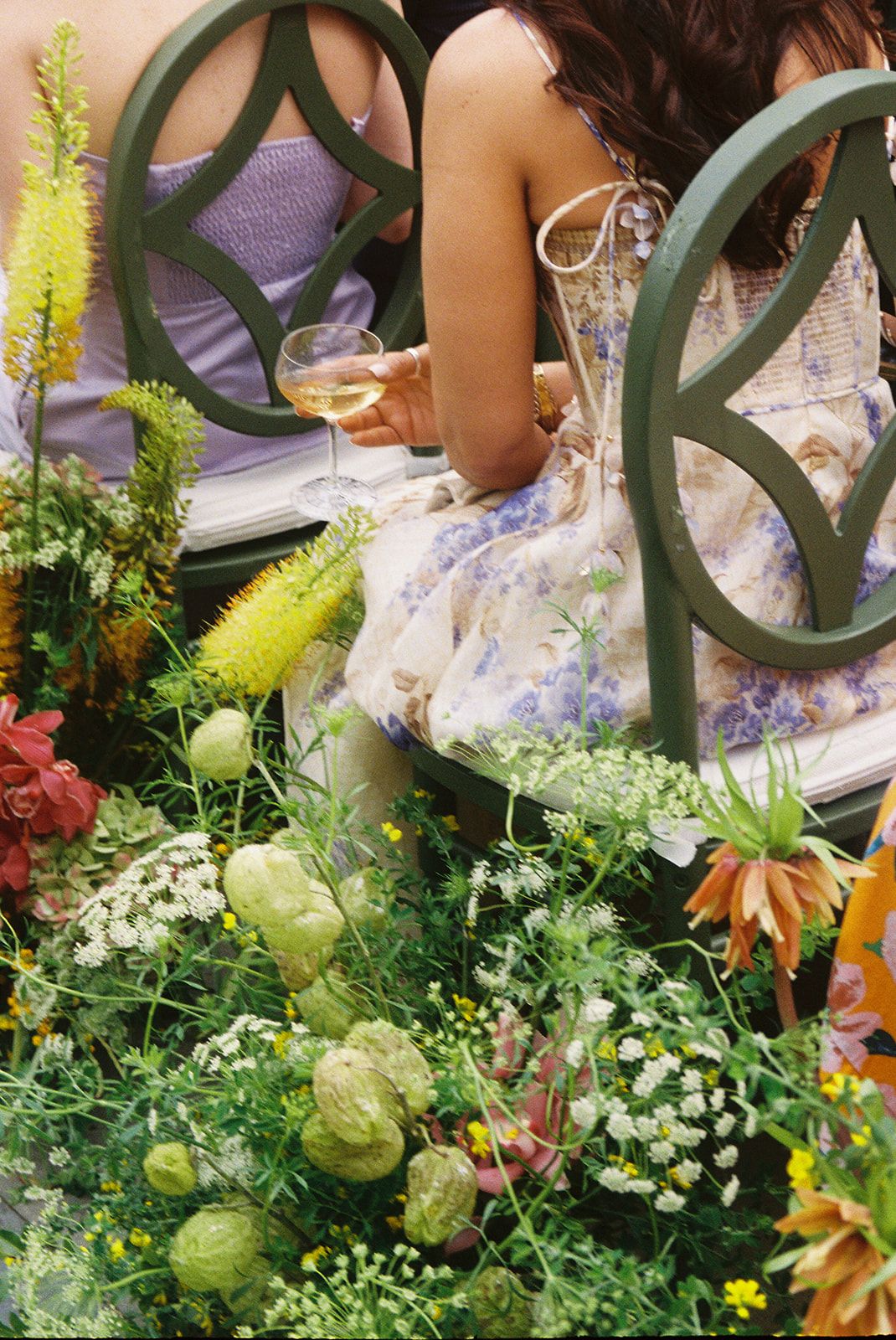 Floral garden party setting with ornate wrought iron chairs and delicate purple and white dress details among lush greenery.