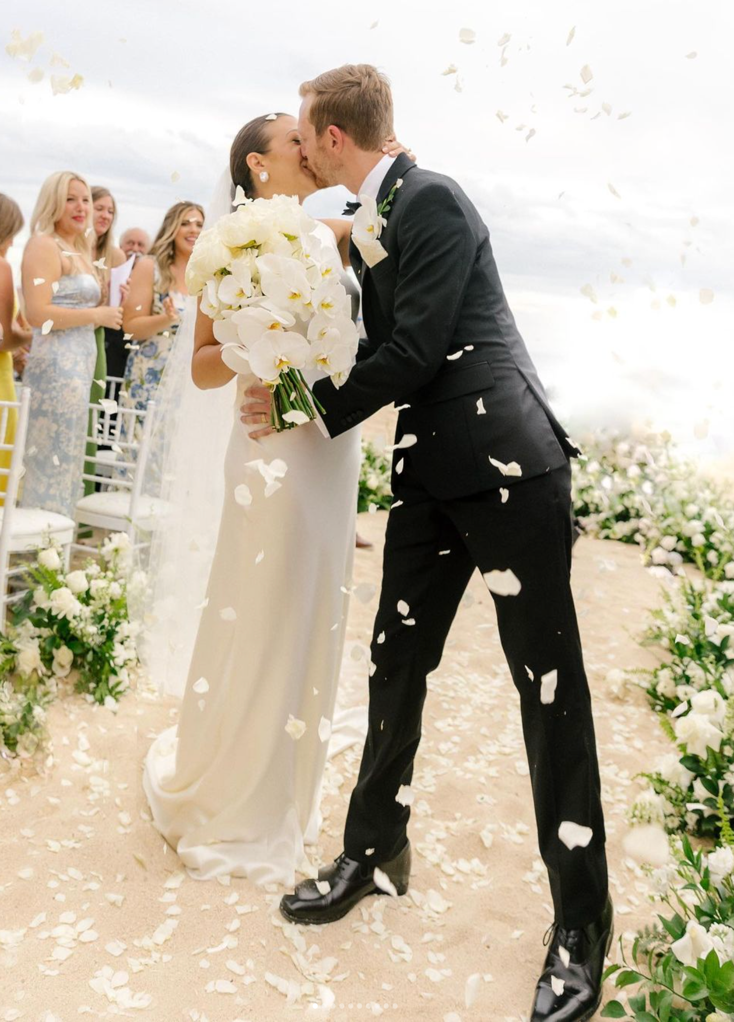 A romantic wedding couple share a kiss outdoors as white flower petals fall around them with guests watching in the background.